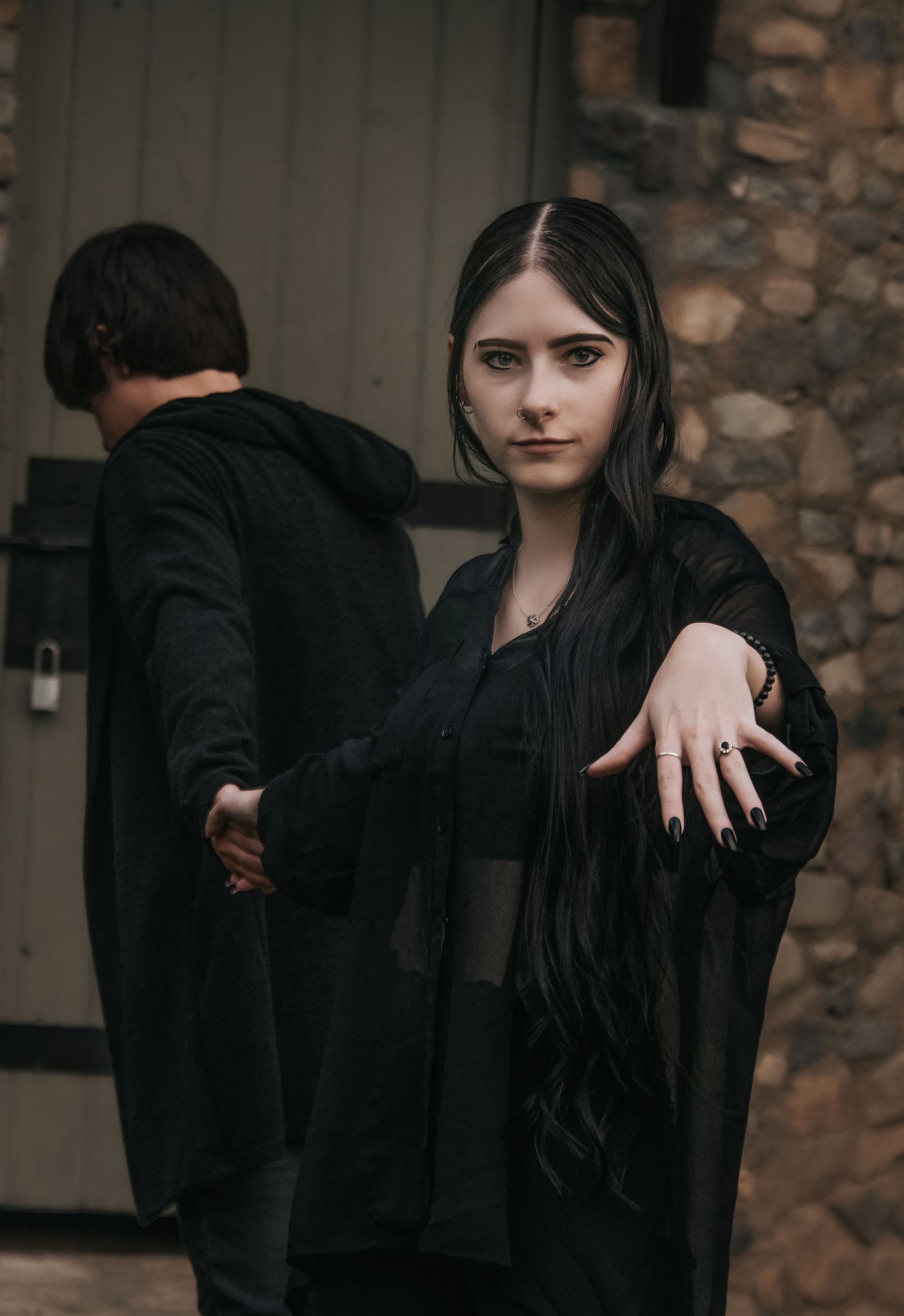 A moody couple in black clothing poses for a dramatic photoshoot against a brick wall.