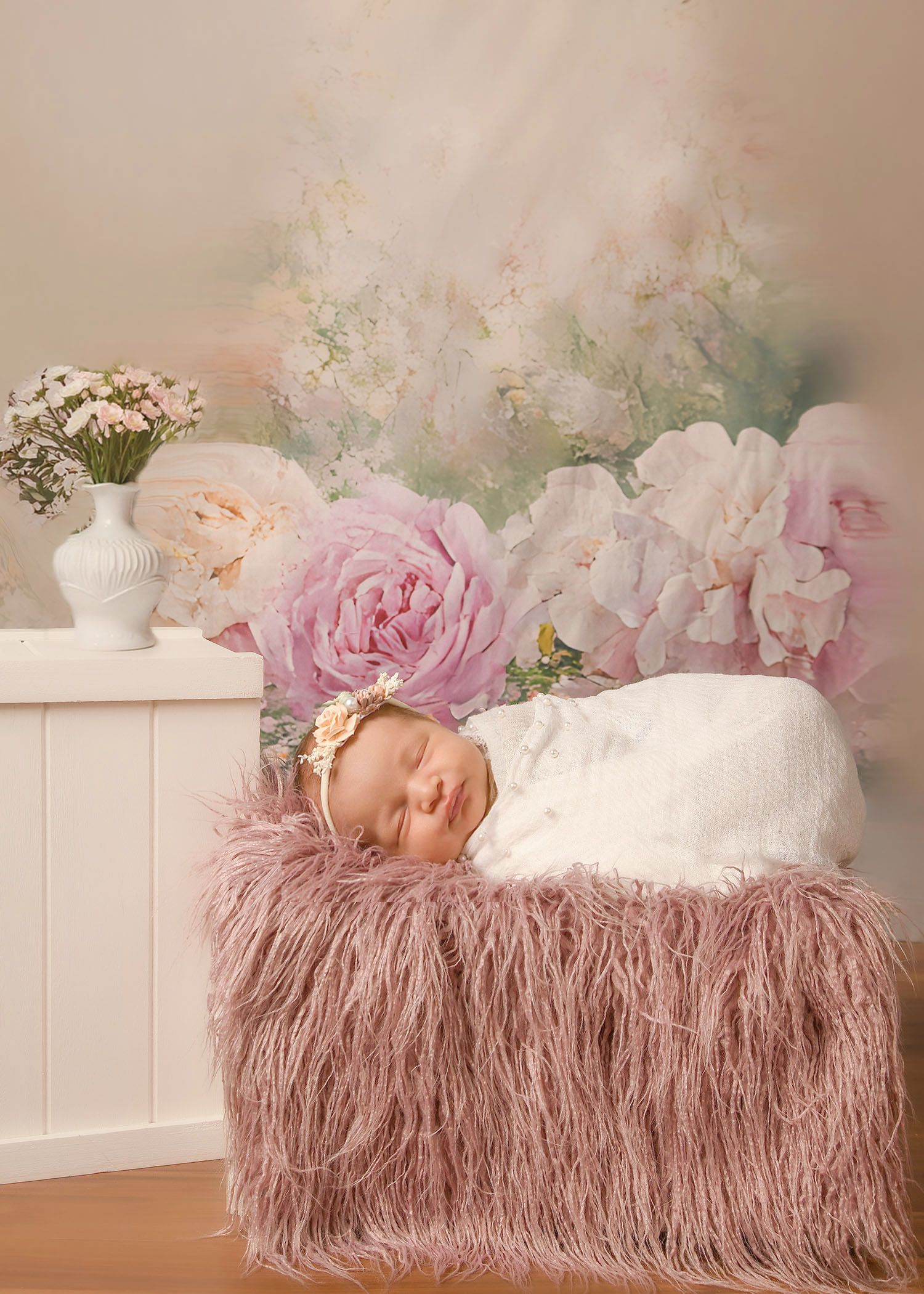 Sleeping newborn baby swaddled in white wrap with floral headband resting on pink fur in front of floral backdrop.