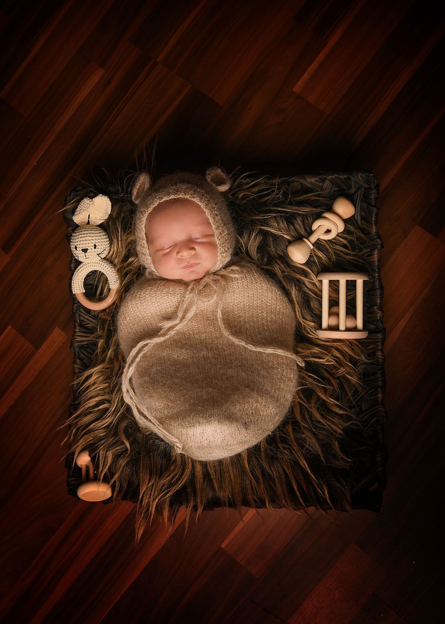 Newborn baby swaddled in brown knit wrap and bear bonnet, sleeping on fur blanket surrounded by wooden toys.