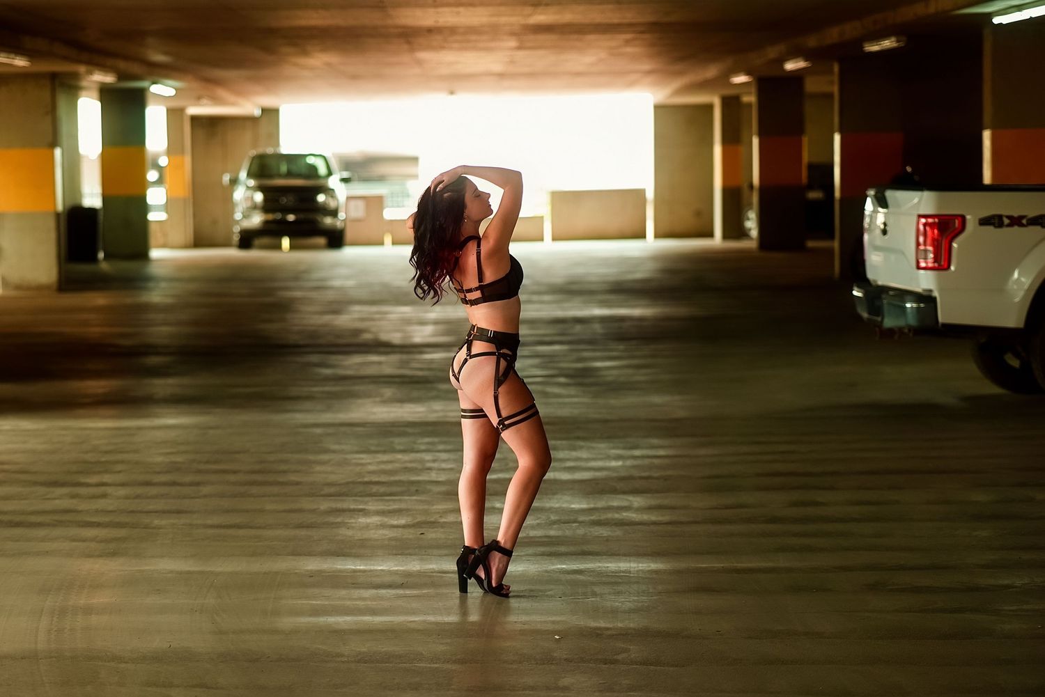 Woman in black lingerie posing in heels inside a dimly lit parking garage with vehicles in the background.