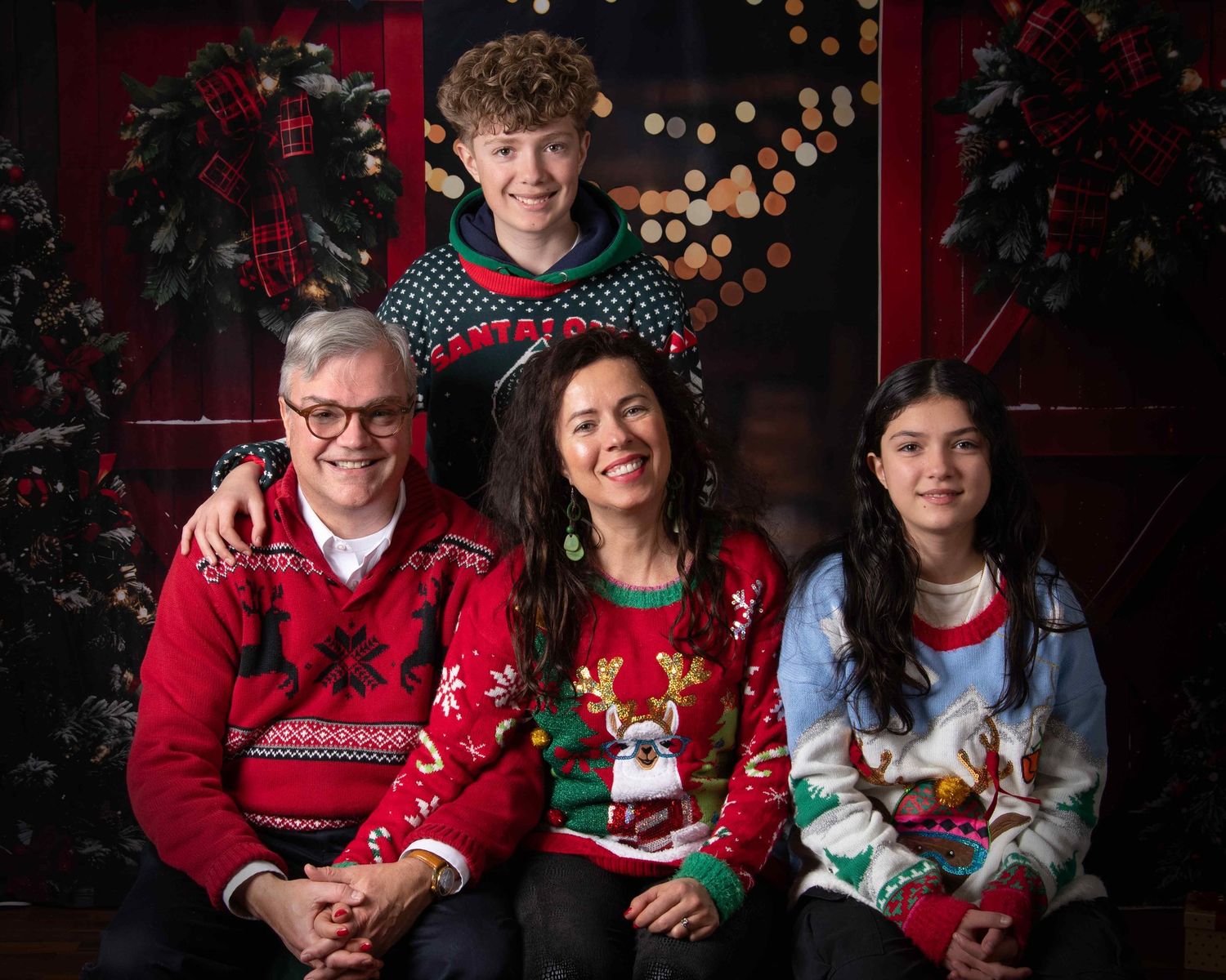 A family wearing festive Christmas sweaters poses together against a dark background with holiday decorations.