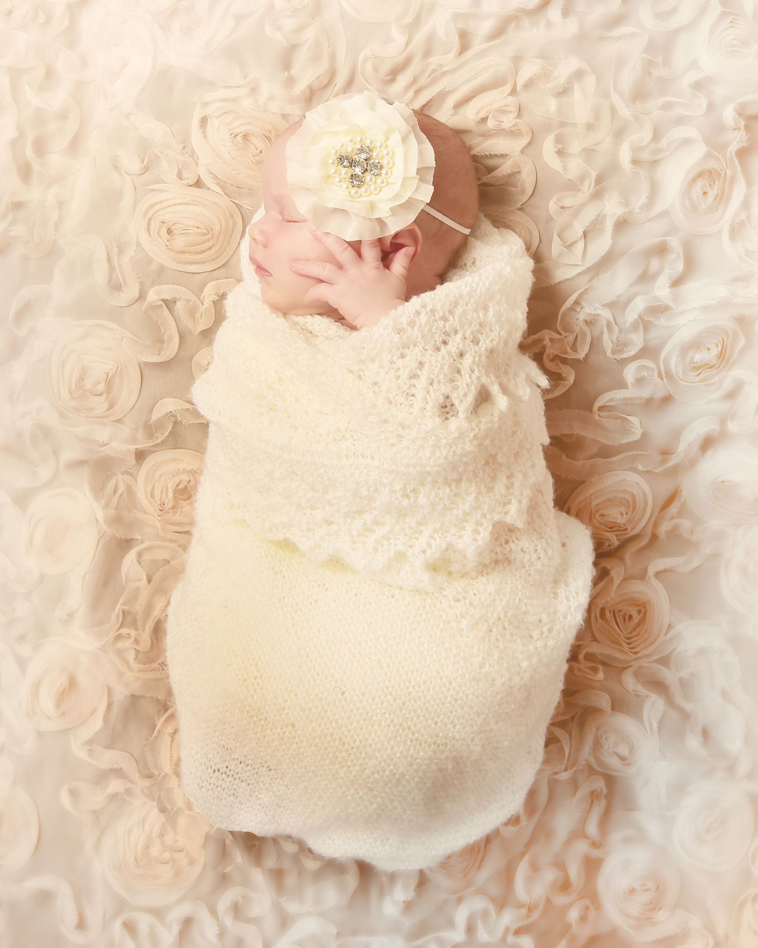 Newborn baby girl swaddled in cream knit wrap with floral headband, lying on blush rosette backdrop.