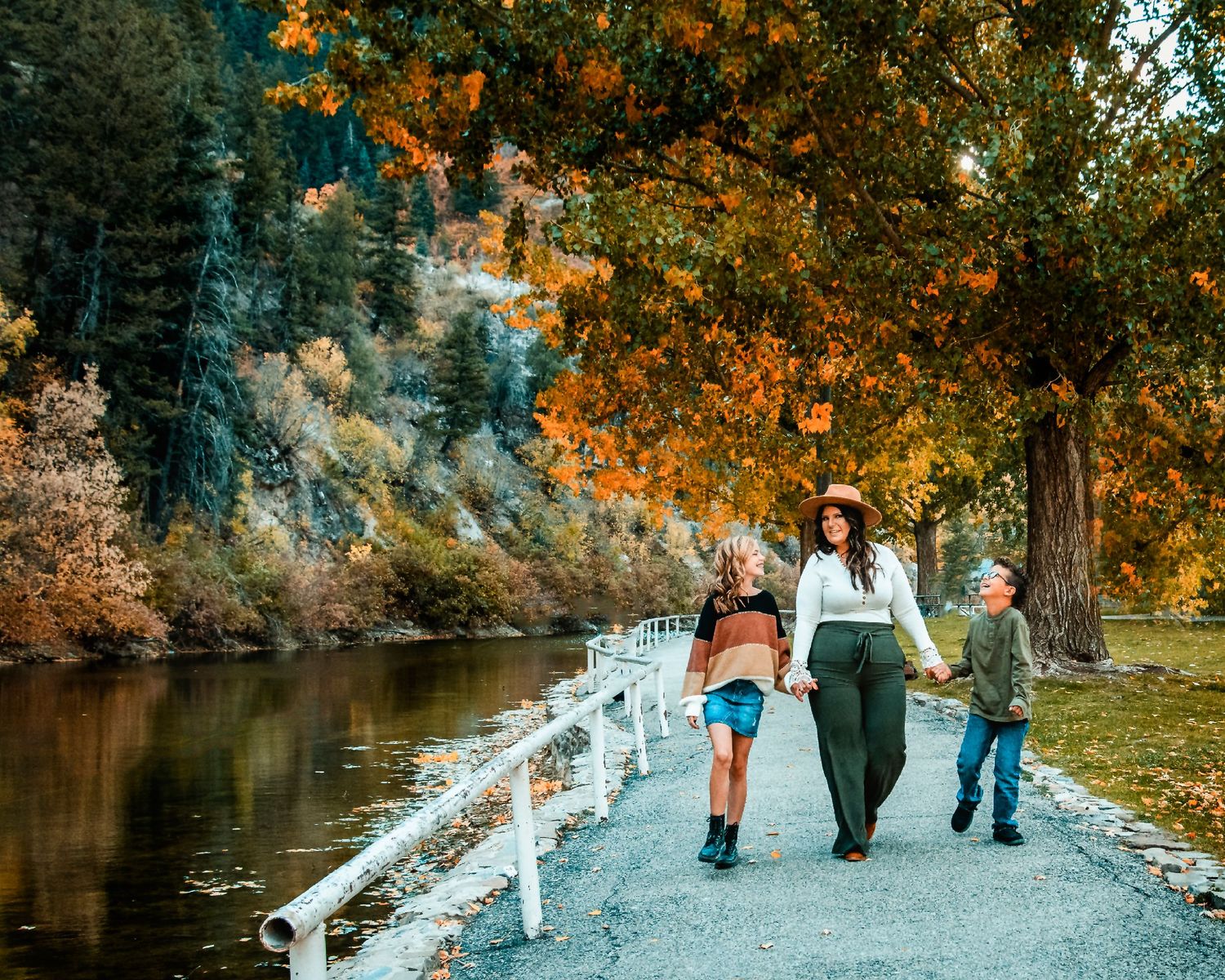 People walking along a riverside path during autumn with colorful foliage overhead.