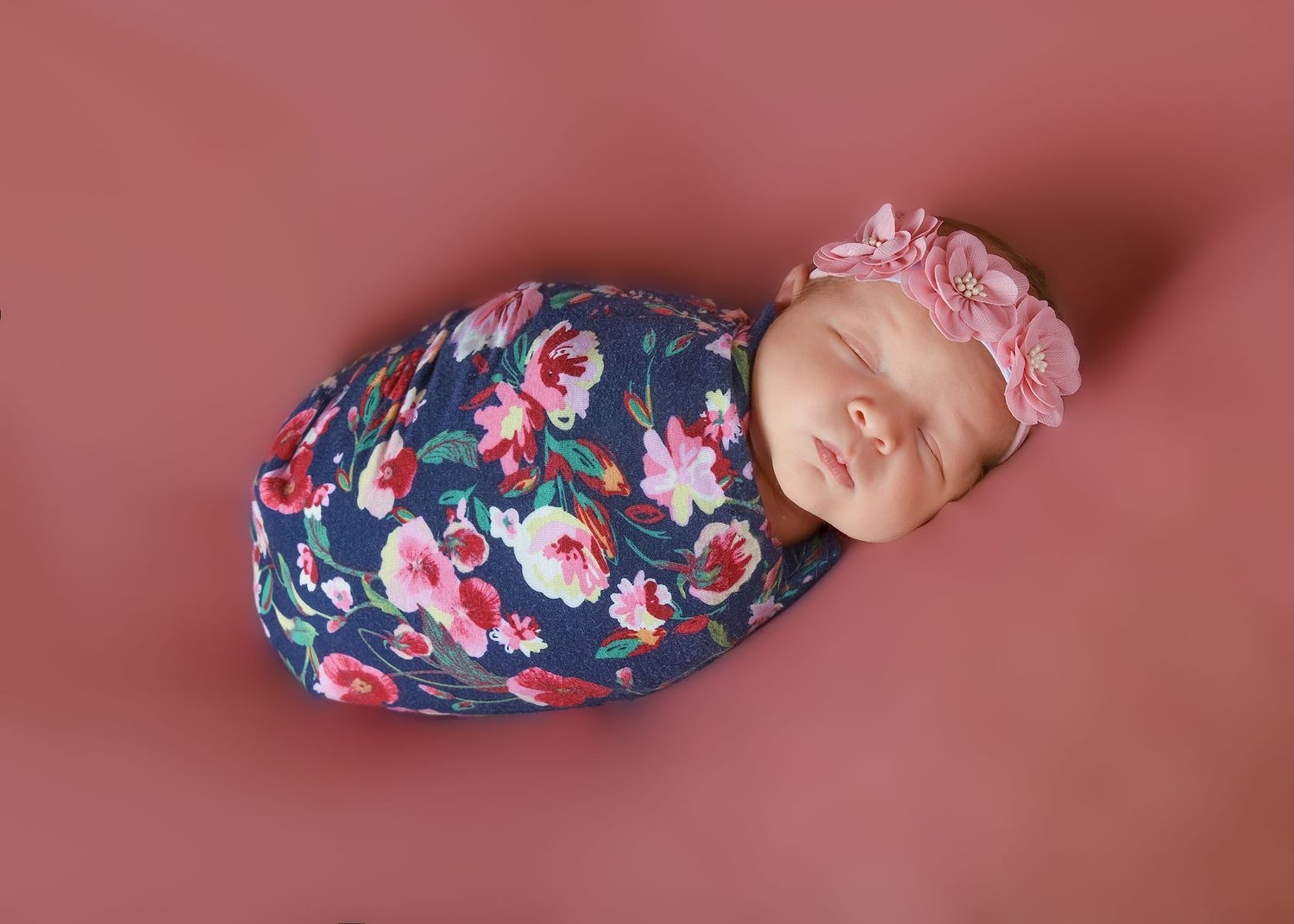 Newborn baby girl swaddled in navy floral wrap with pink rose headband, sleeping peacefully on pink background.