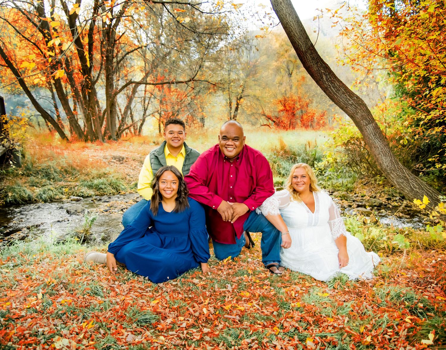 Group sitting on fallen autumn leaves near a stream with colorful fall trees.