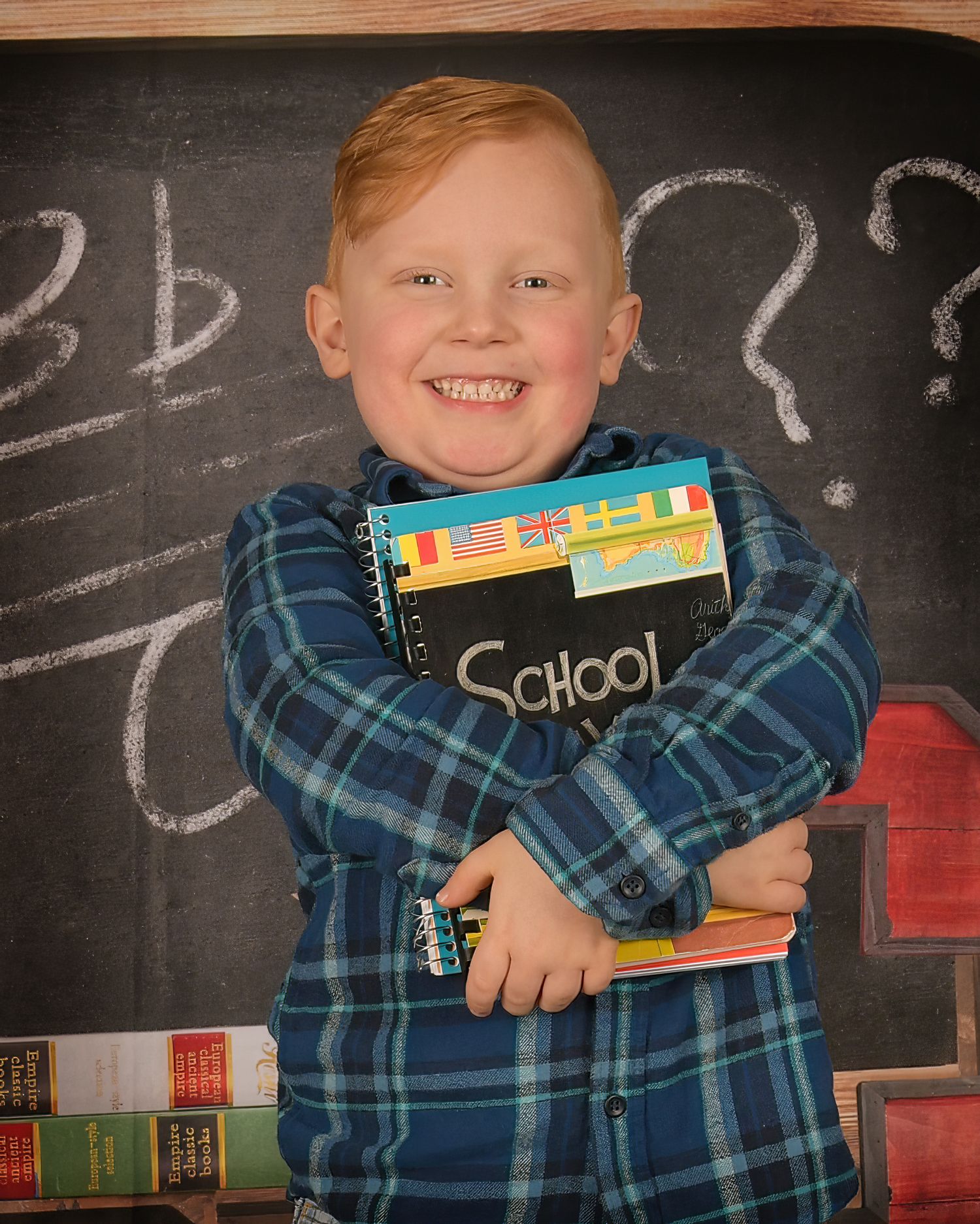 A student wearing a plaid shirt hugs school supplies while standing in front of a chalkboard with question marks drawn on it.