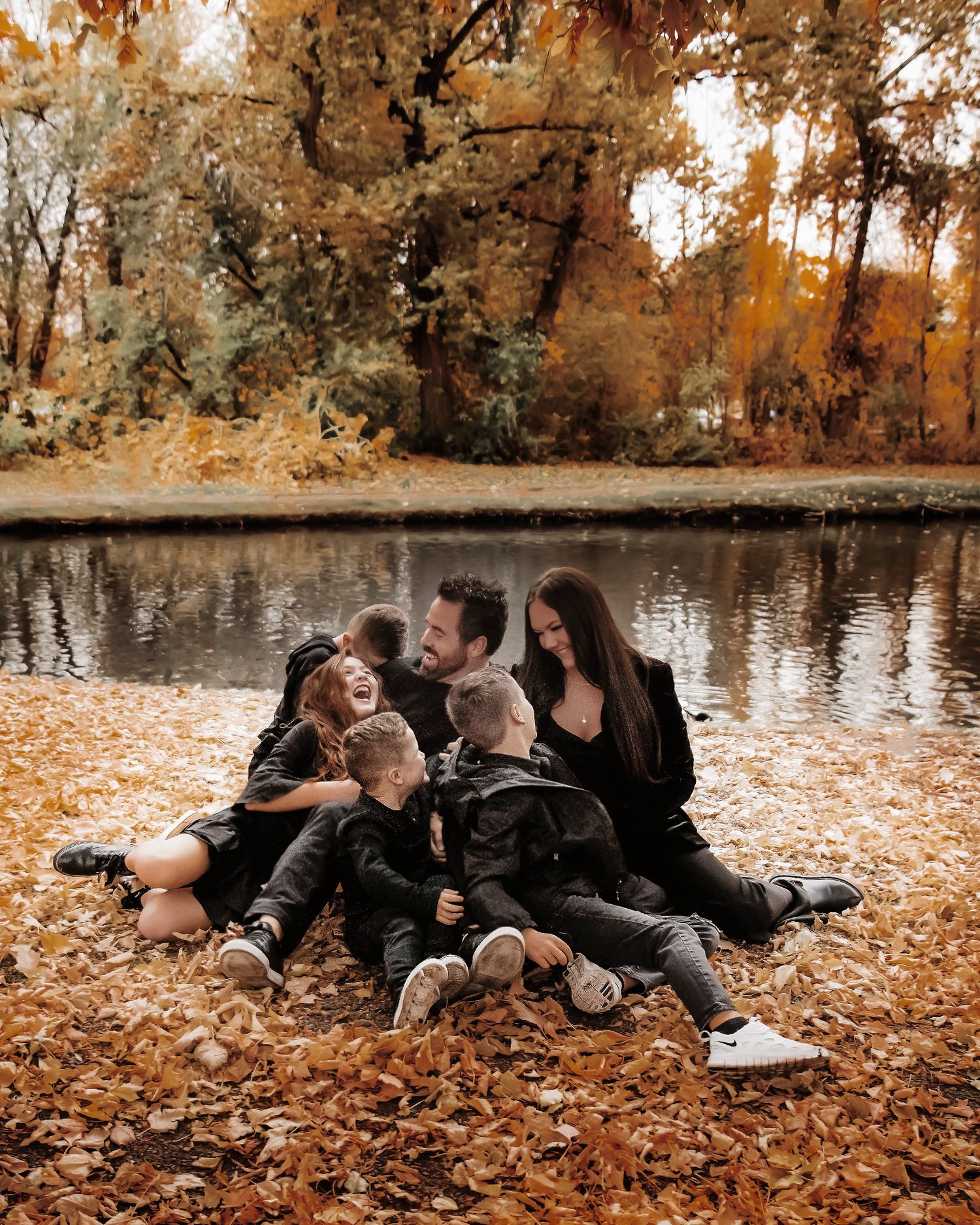 Family relaxing together on fallen autumn leaves beside a tranquil lake during a fall photo session.