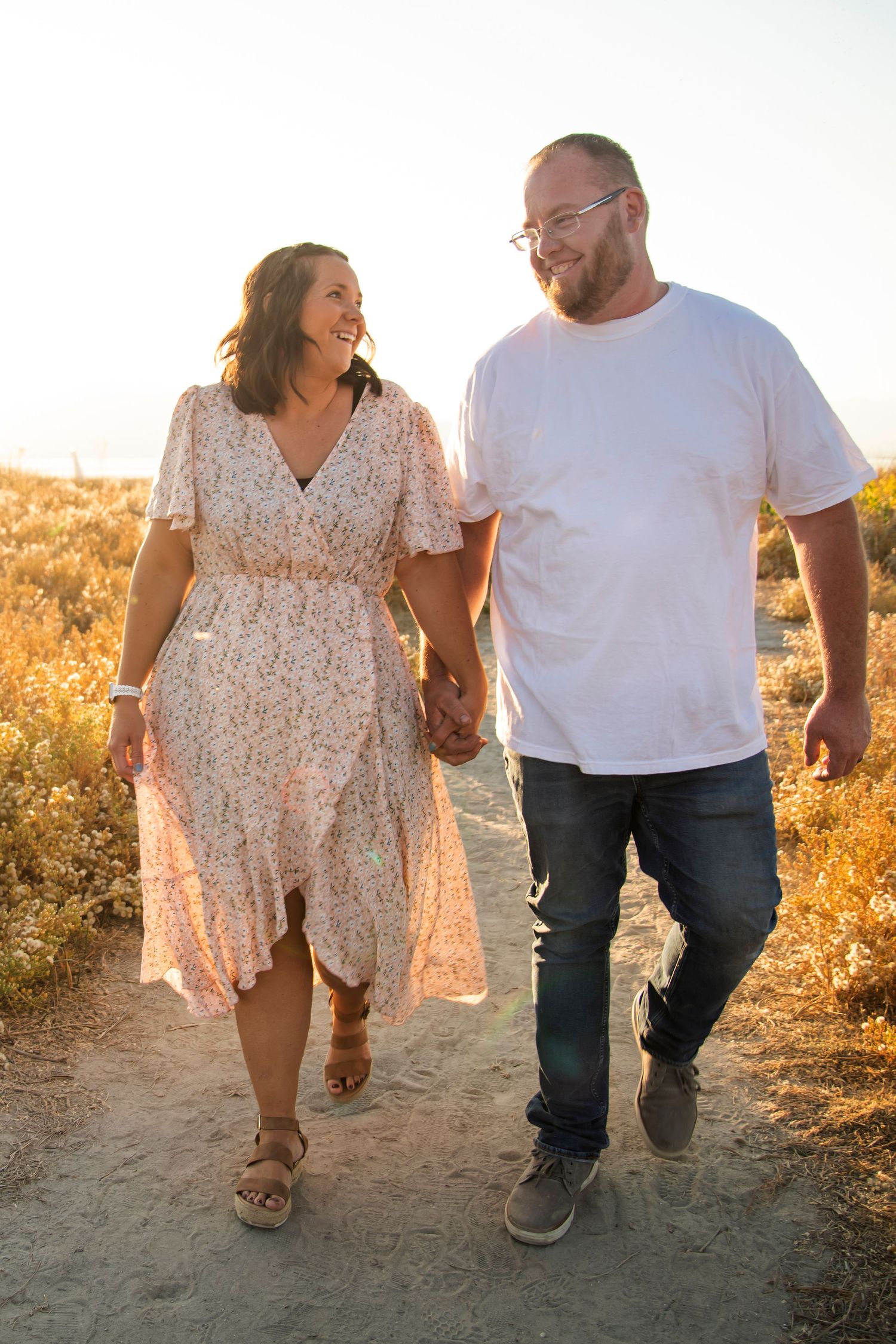 Couple walking hand in hand on dirt path through golden field at sunset.
