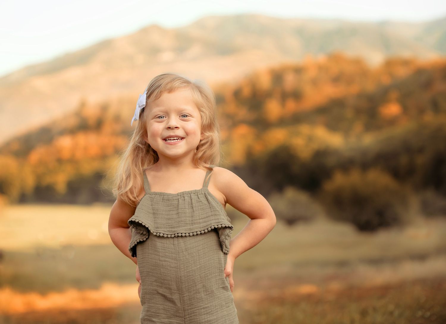 Young child in gray dress poses confidently against backdrop of autumn mountains.