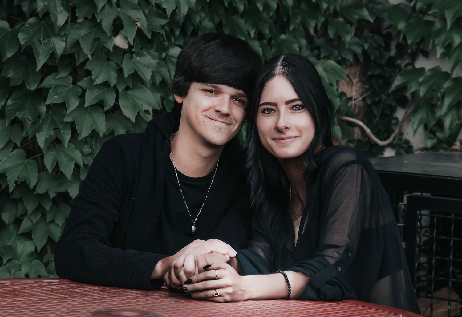 A couple wearing black outfits sits together in a garden setting with lush green ivy in the background.