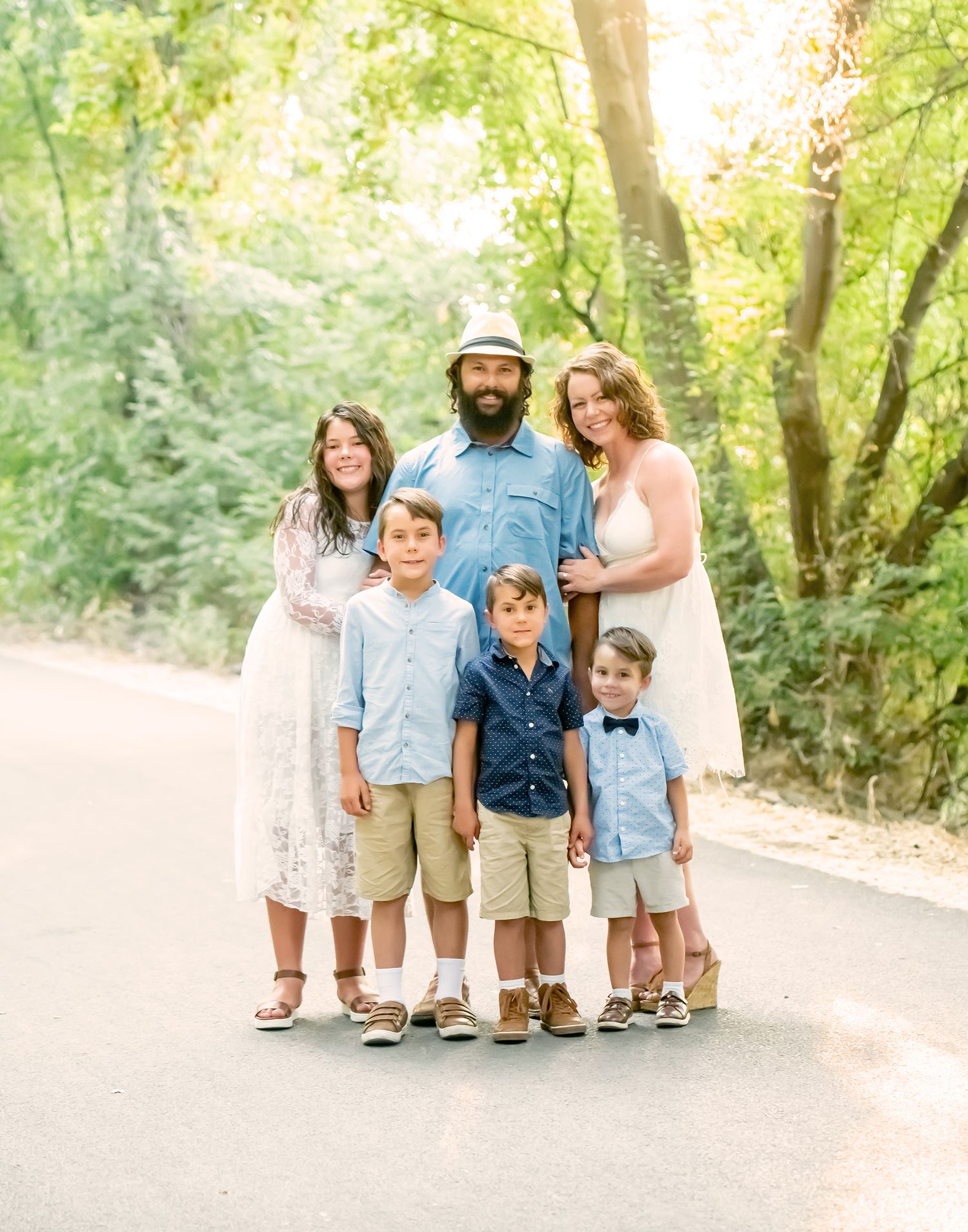 Family group portrait on a sunlit forest path wearing coordinated light-colored outfits.