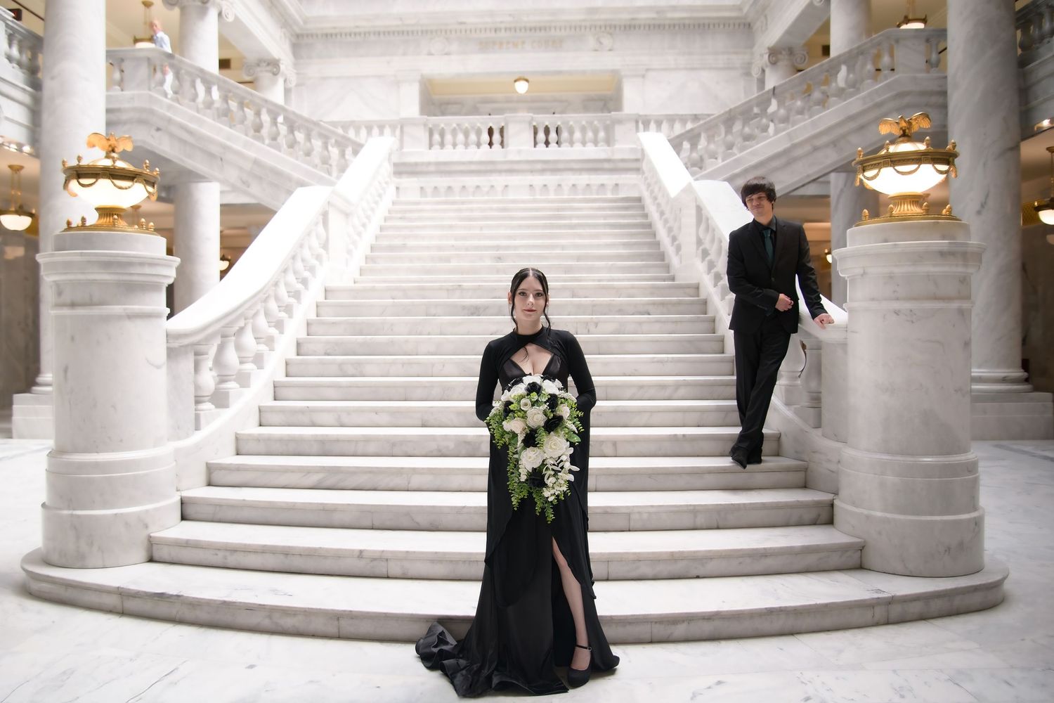 Gothic wedding couple posed on an elegant white marble staircase in an ornate building.