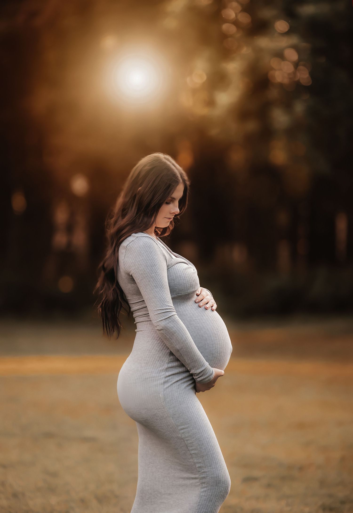 A pregnant woman in a fitted gray dress stands in profile against a warm sunset backdrop in a natural outdoor setting.