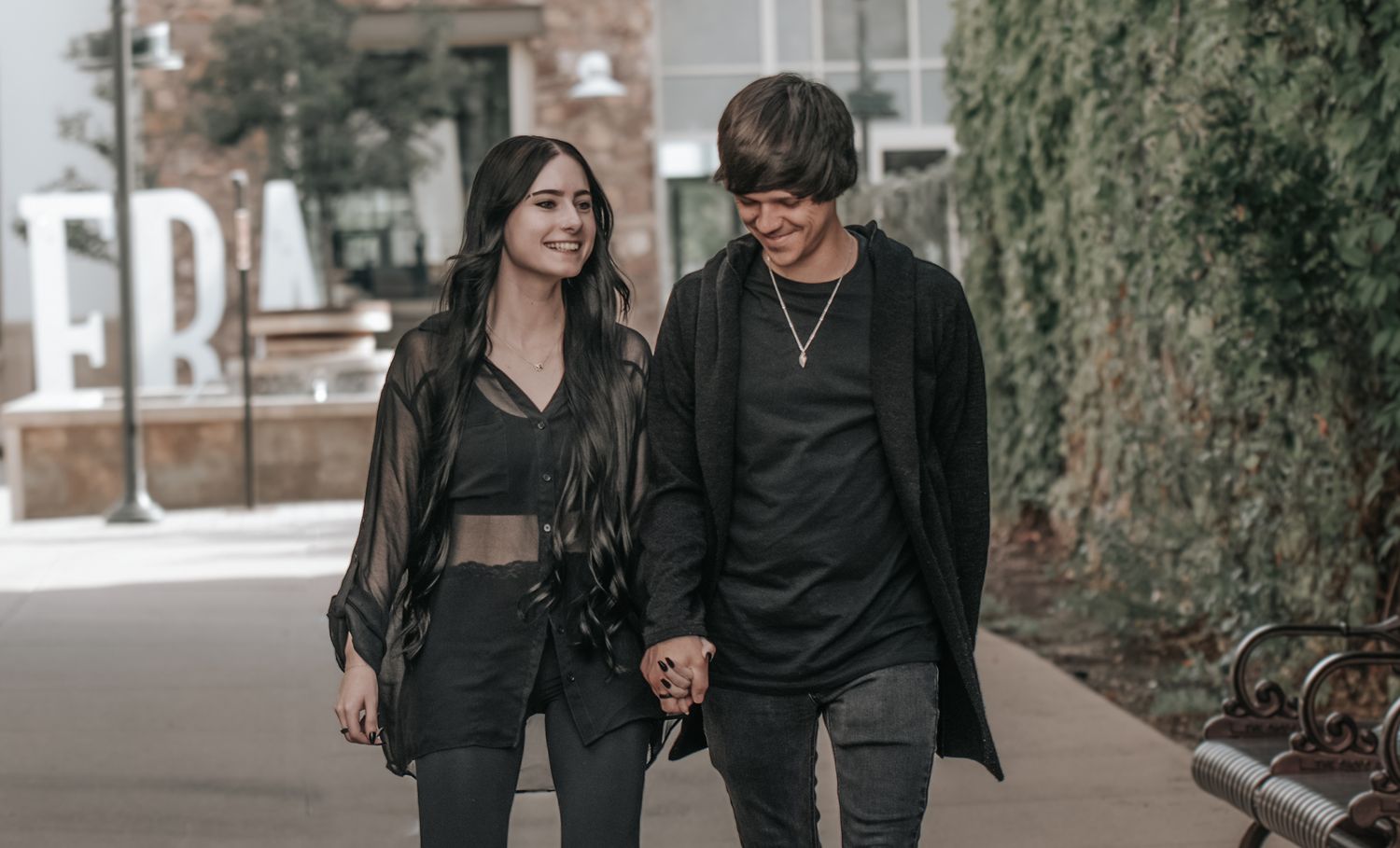 A couple in matching black outfits walks together on an urban path while holding hands.