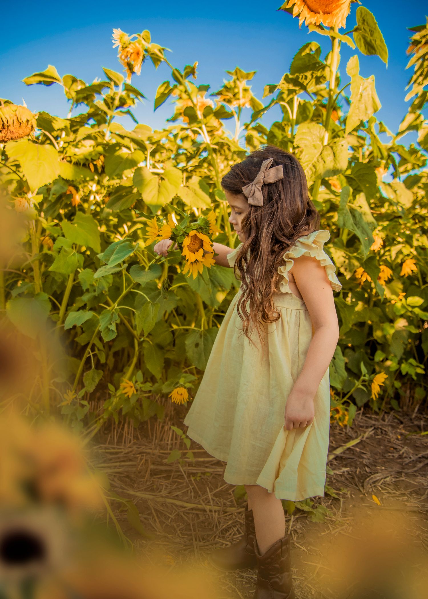 A girl in a yellow dress surrounded by towering sunflowers against a bright blue sky.