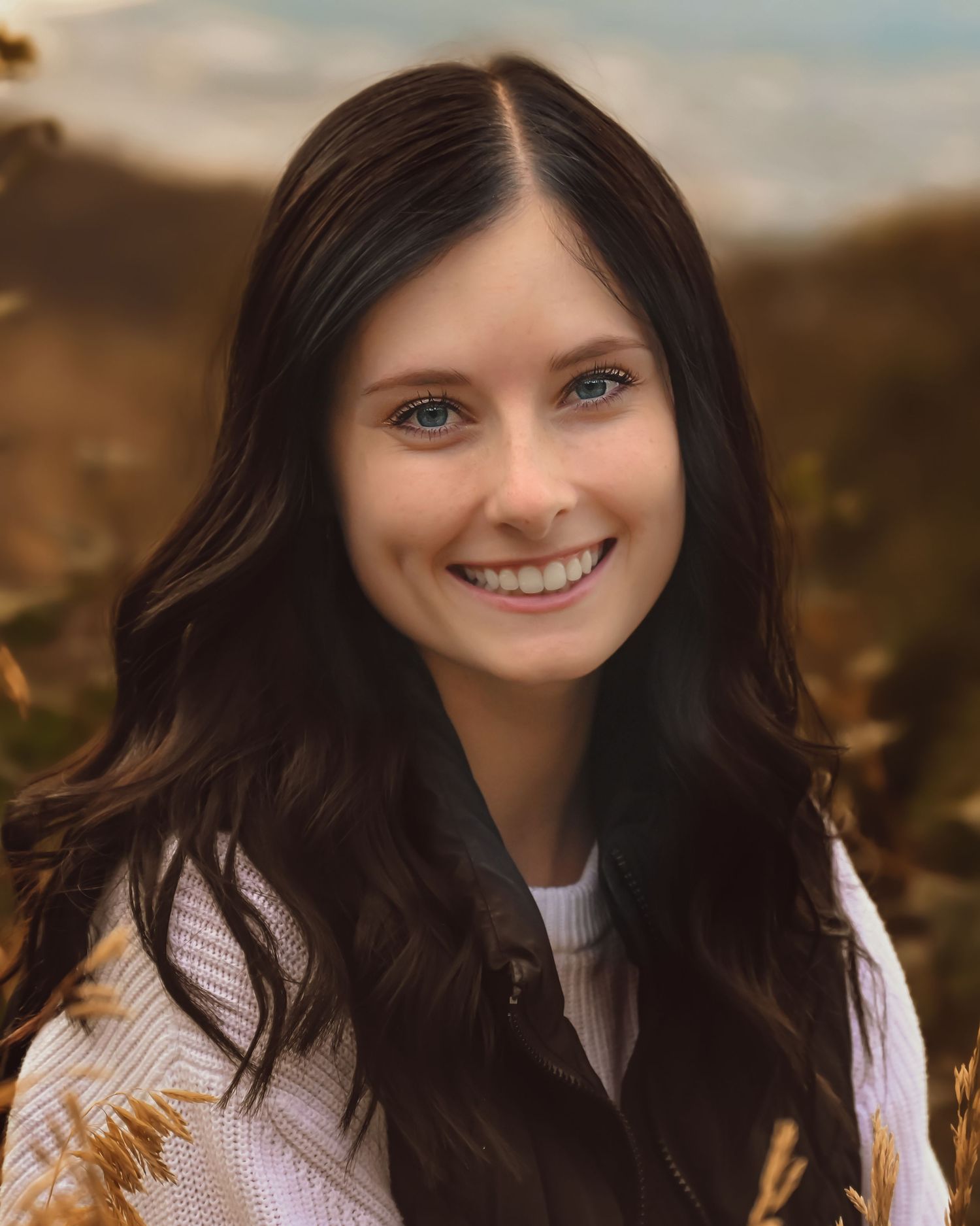 Portrait of a woman with long dark hair wearing a white sweater against an outdoor autumn backdrop.
