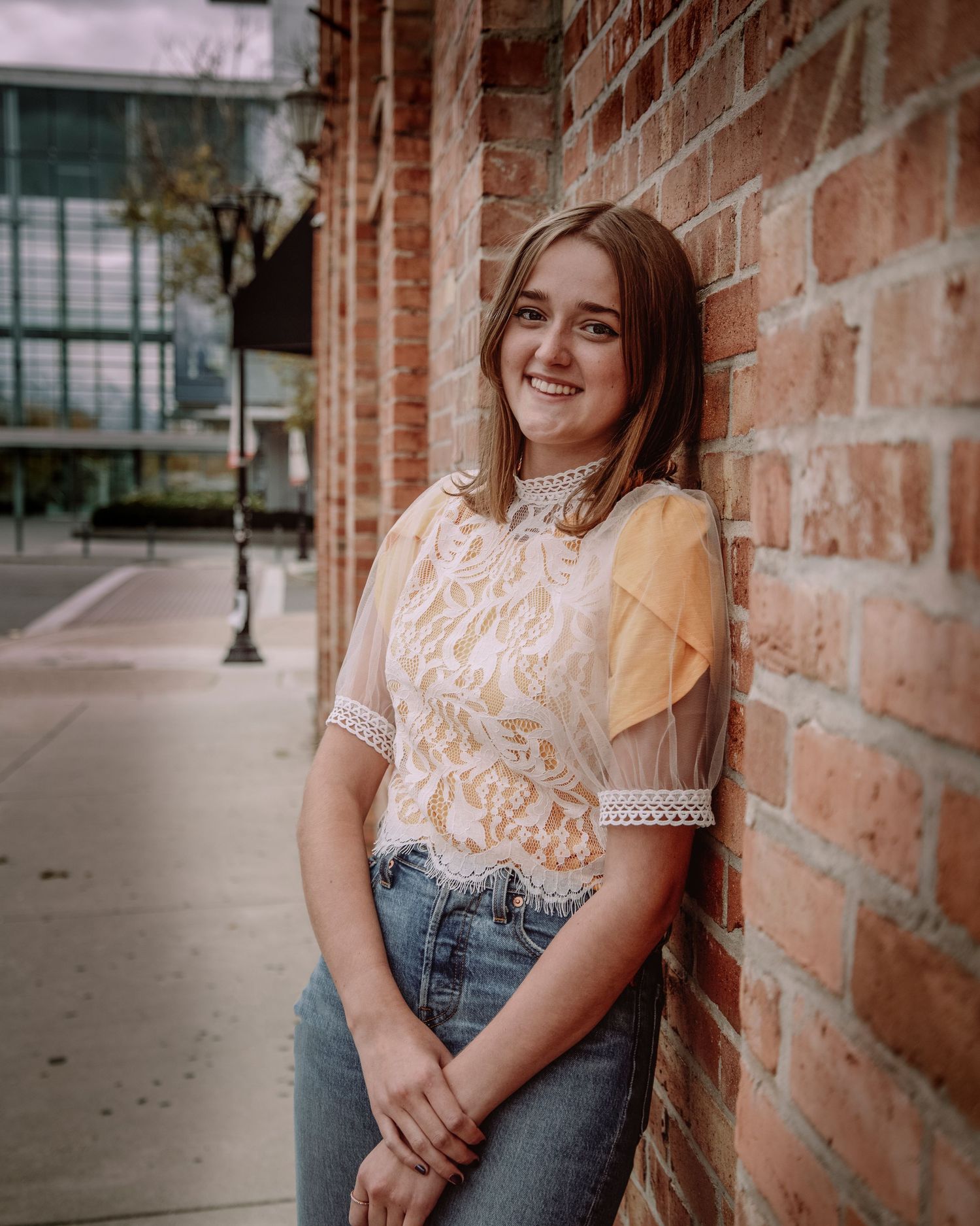 Young woman in a lace top and jeans smiles while leaning against a brick wall.