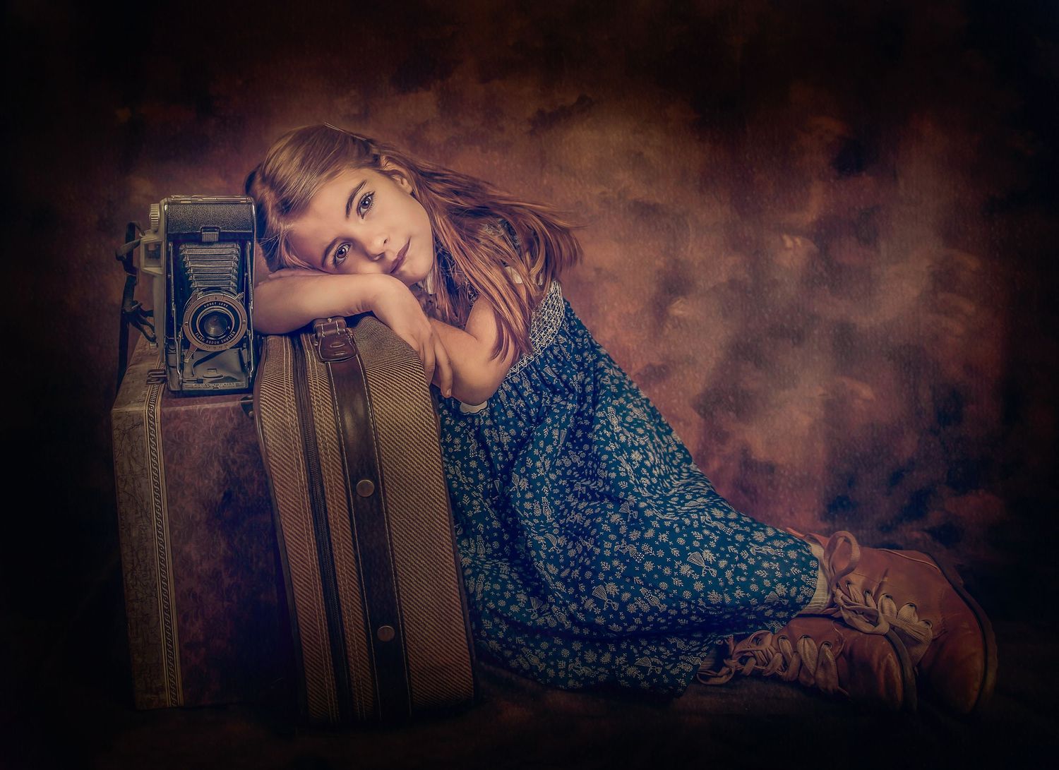 A whimsical portrait of a child resting on vintage luggage in a blue floral dress.