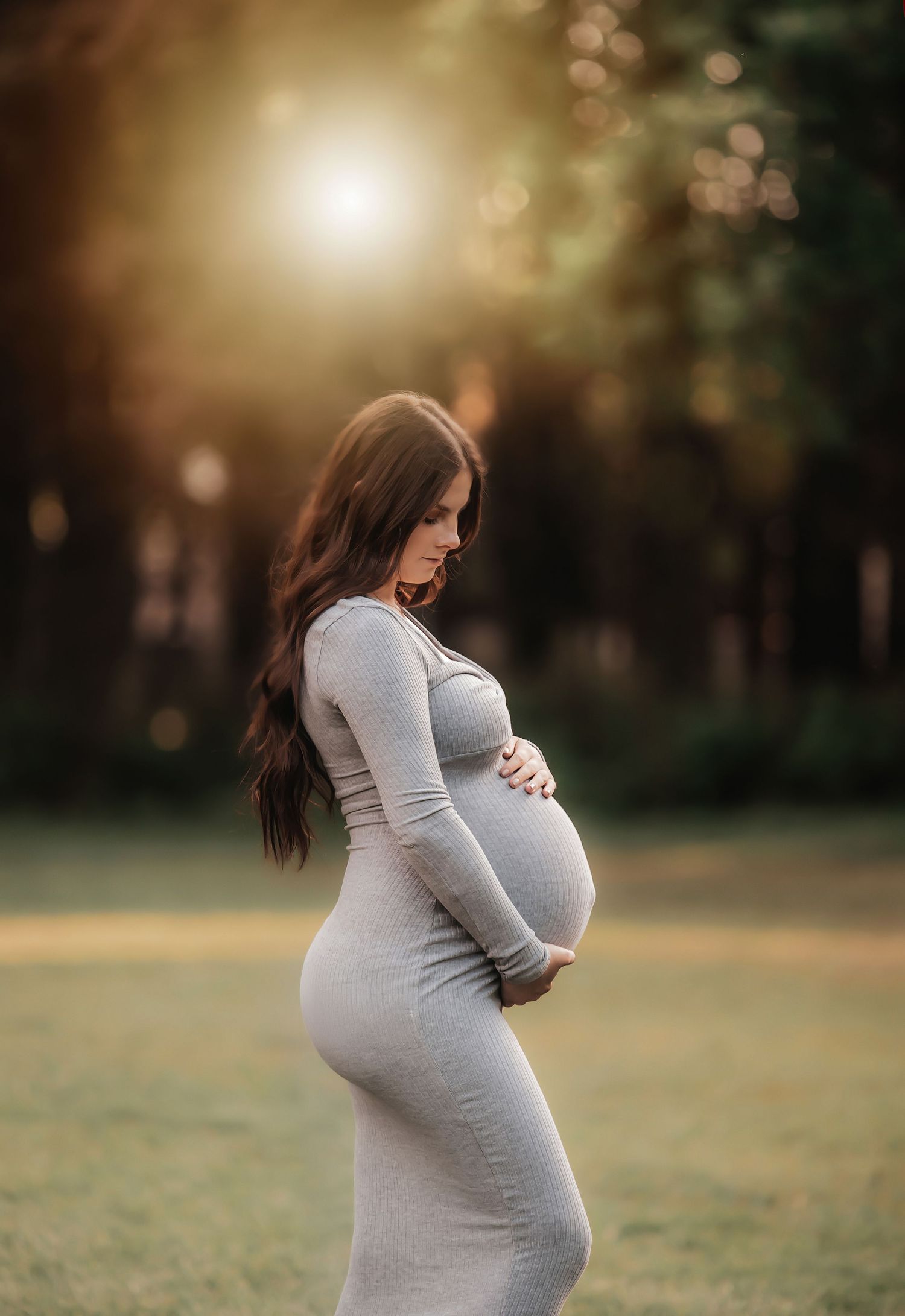 A pregnant woman in a gray dress cradles her baby bump during a golden hour maternity photoshoot in a park.