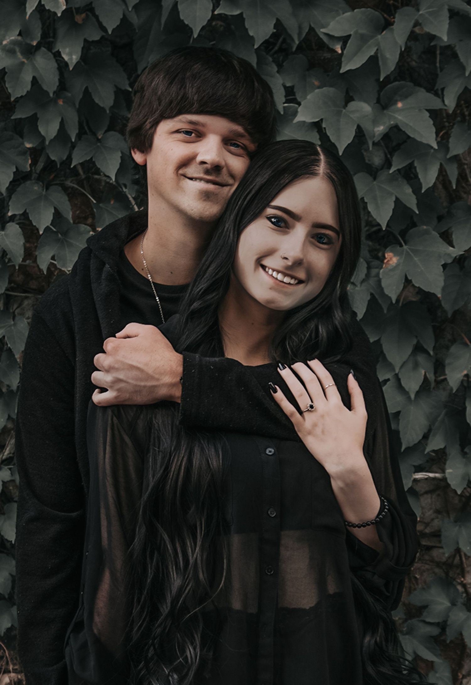 A couple in dark clothing poses against a leafy vine backdrop in an intimate portrait series.