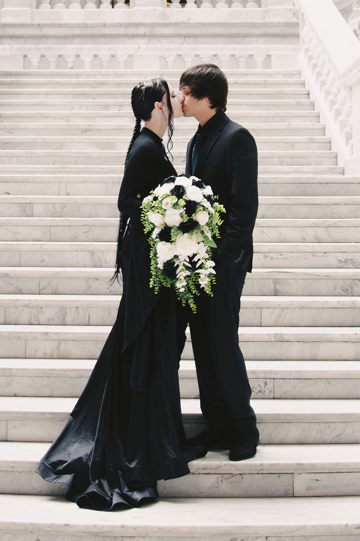A couple in black formal attire share a romantic moment on elegant white stone steps with a white floral bouquet.