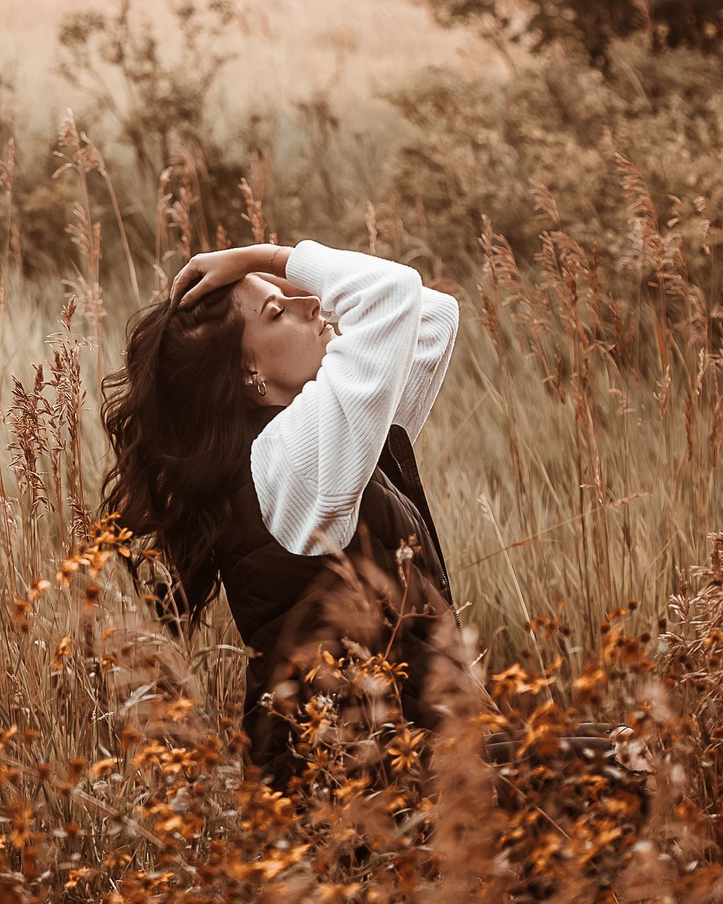 Person in white sweater lying in autumn field with brown grass and dried plants creating moody fall atmosphere.