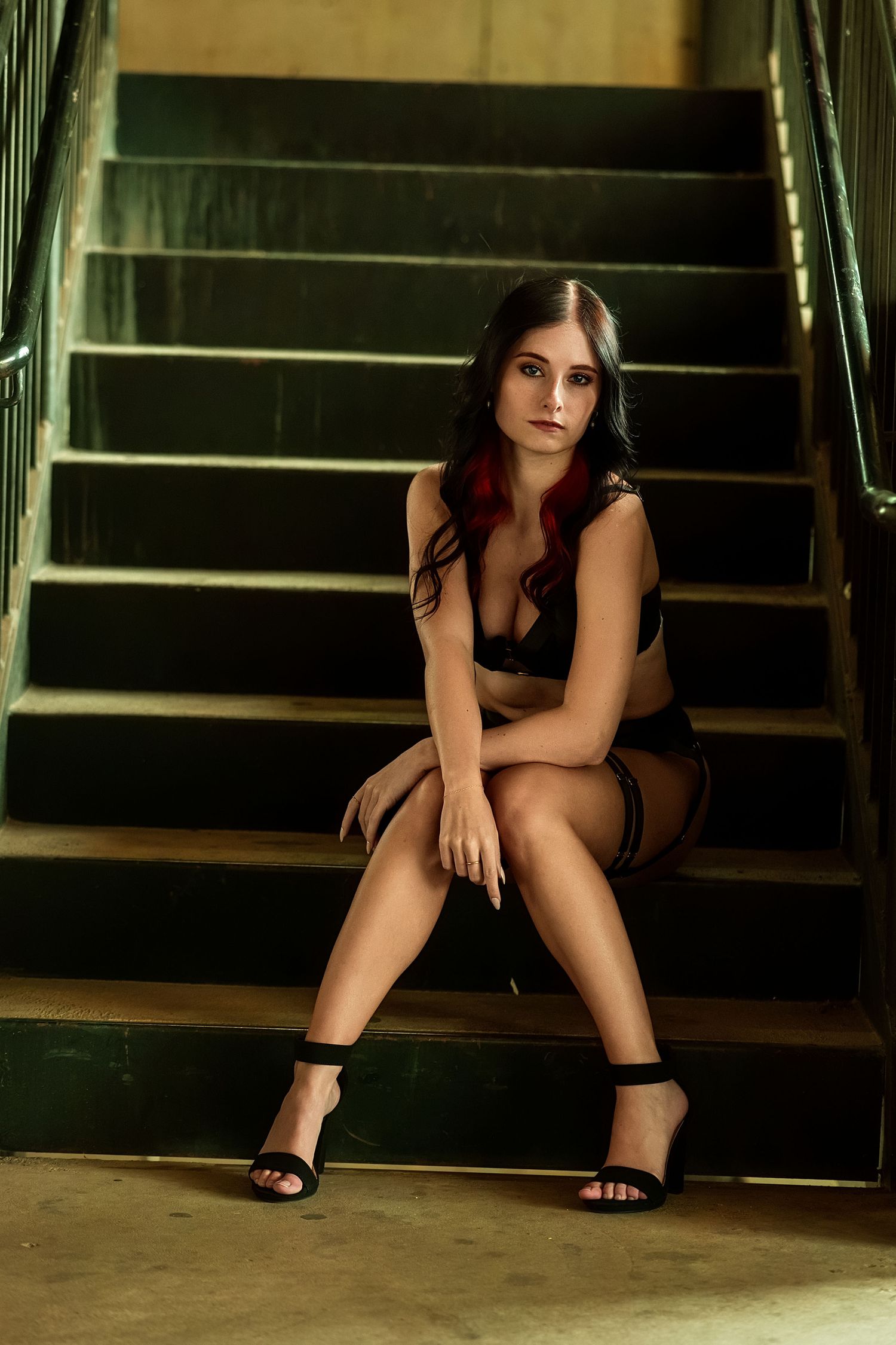 Young woman in black lingerie and heels sitting on dark staircase steps in moody dramatic lighting.