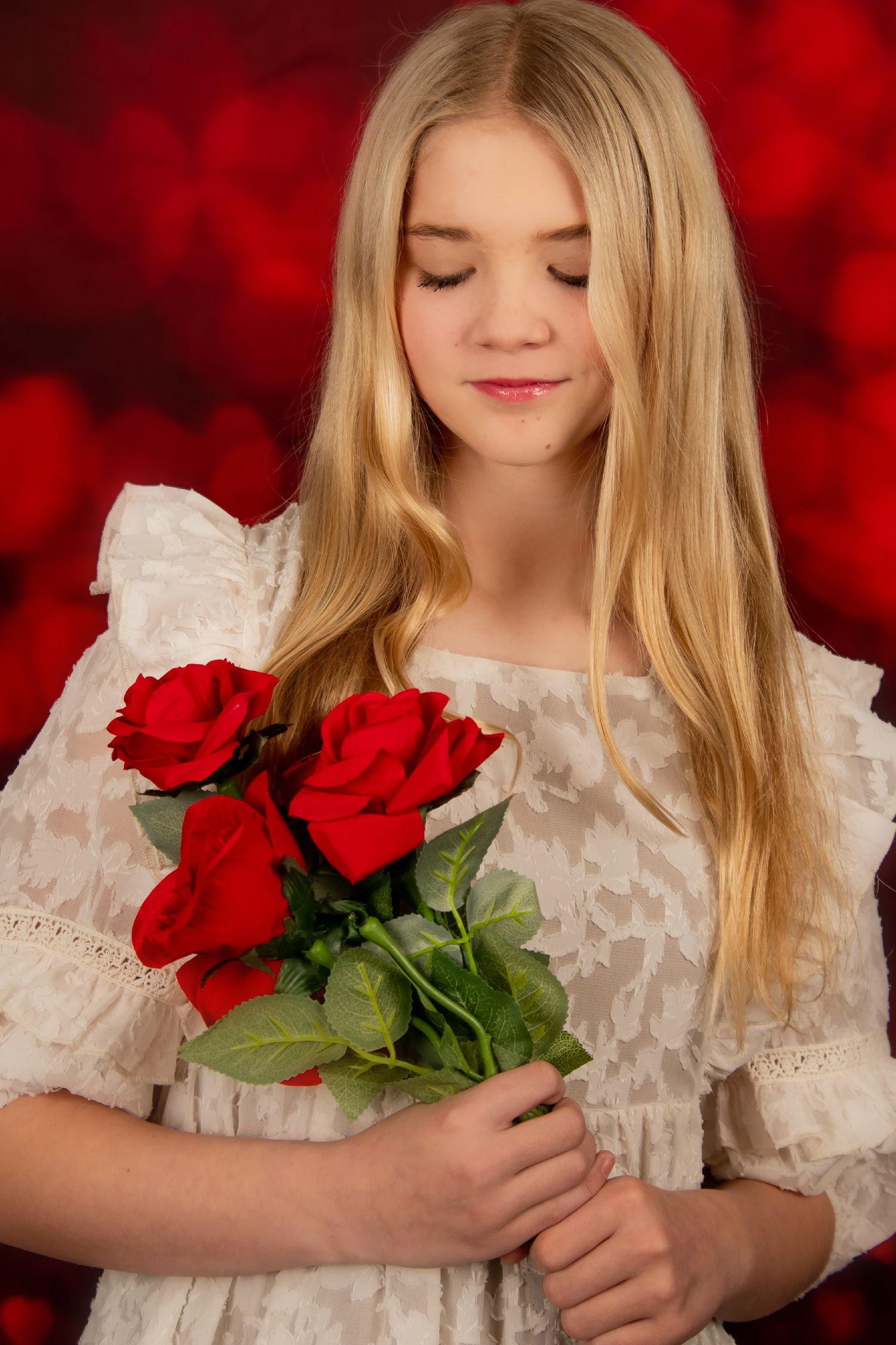 A young person in a white lace dress holds vibrant red roses against a romantic red bokeh background.
