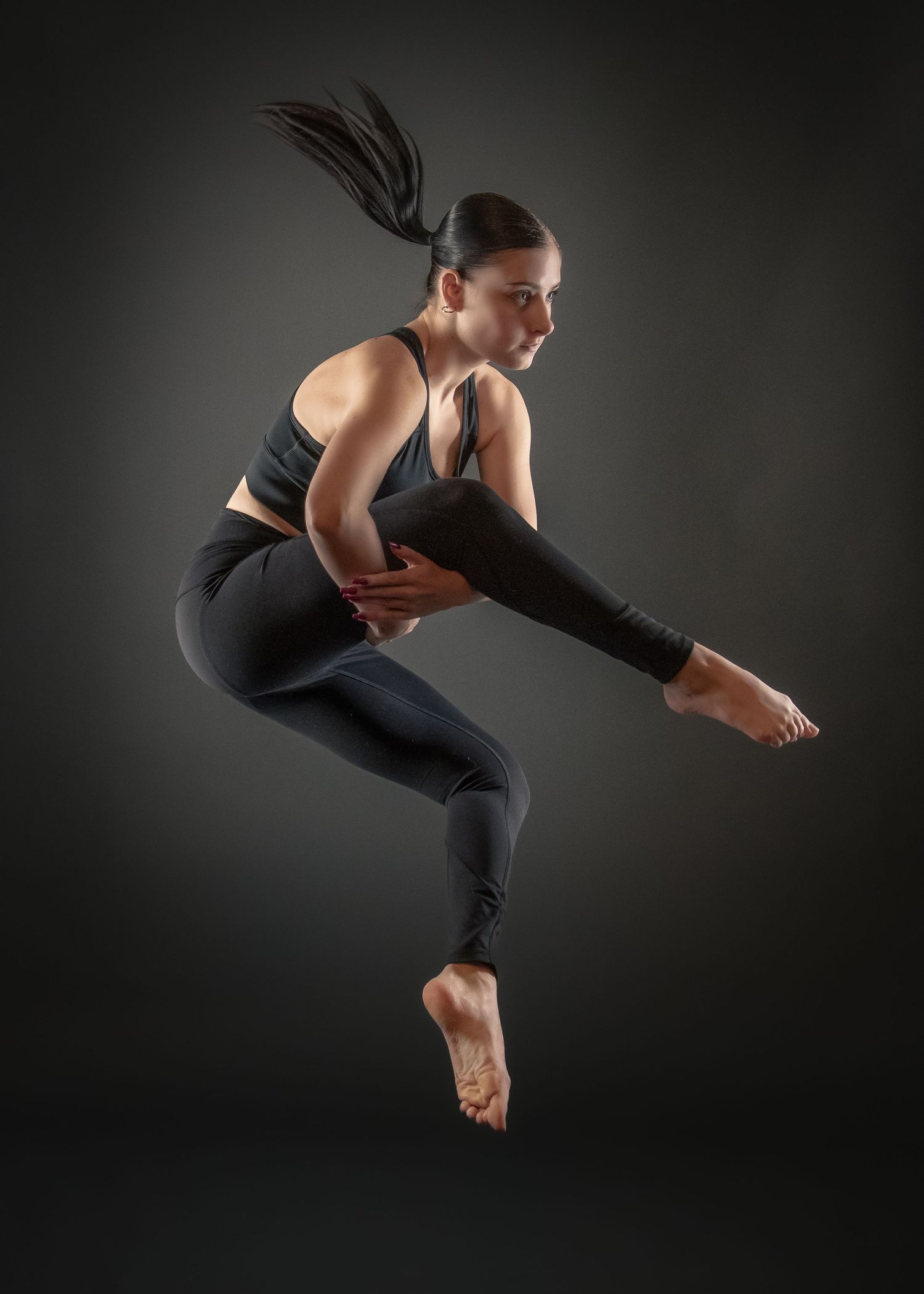 Athletic dancer in black workout attire performs a dynamic leap position against dark background.