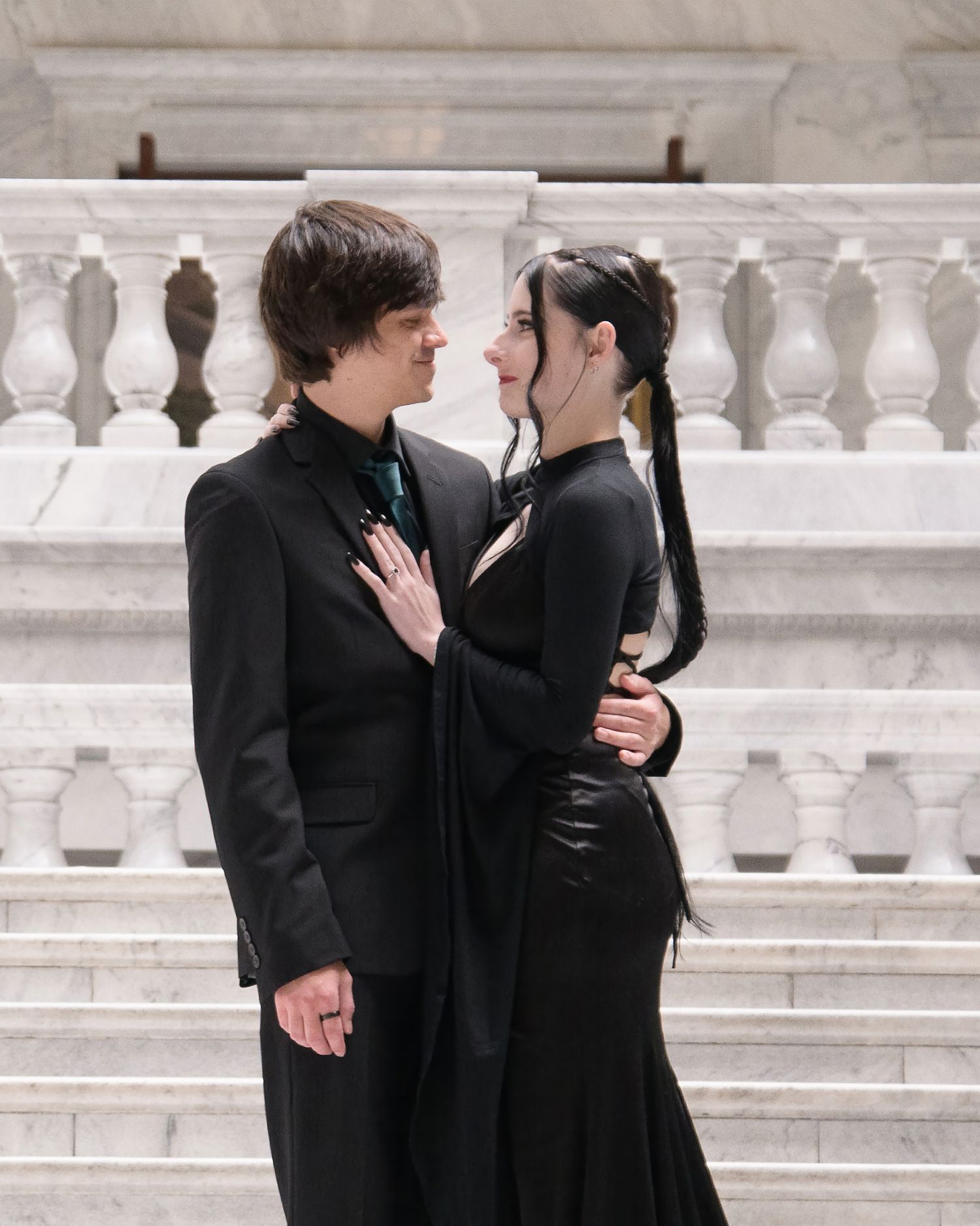Two people in black outfits embrace romantically on vintage marble steps with ornate balustrade railings.