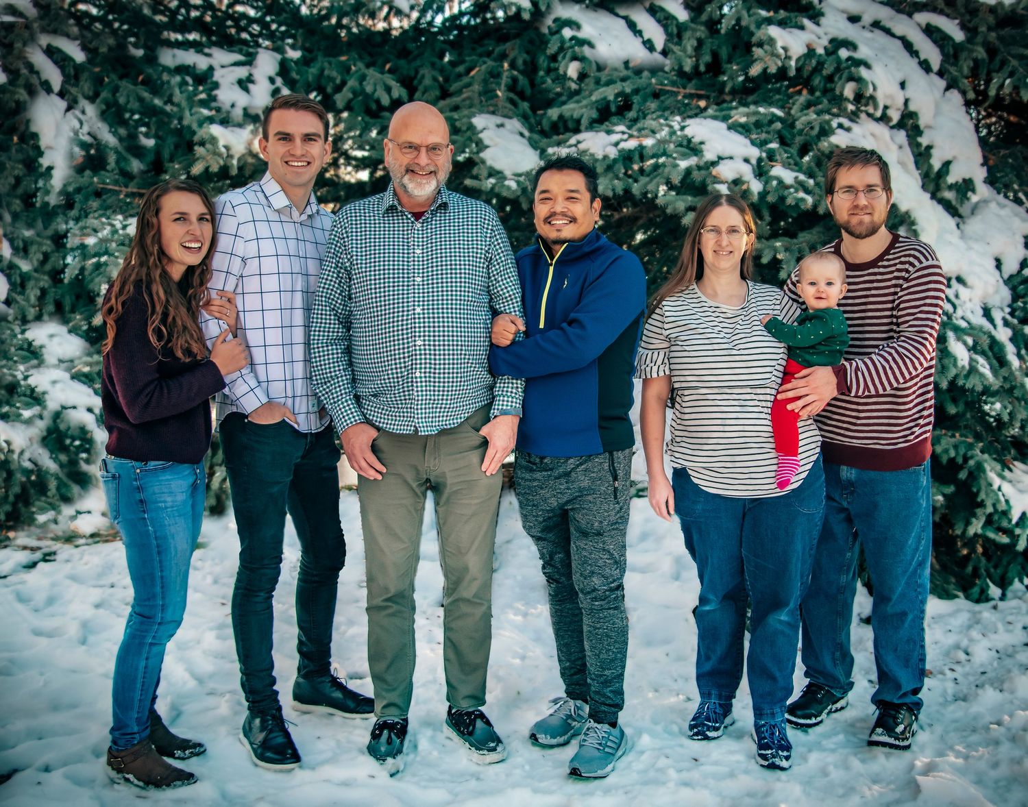 A group photo of six people standing together in the snow in front of evergreen trees.