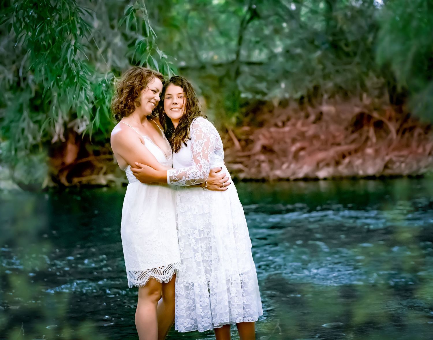 Two people in white dresses embracing by a serene stream in a wooded setting.