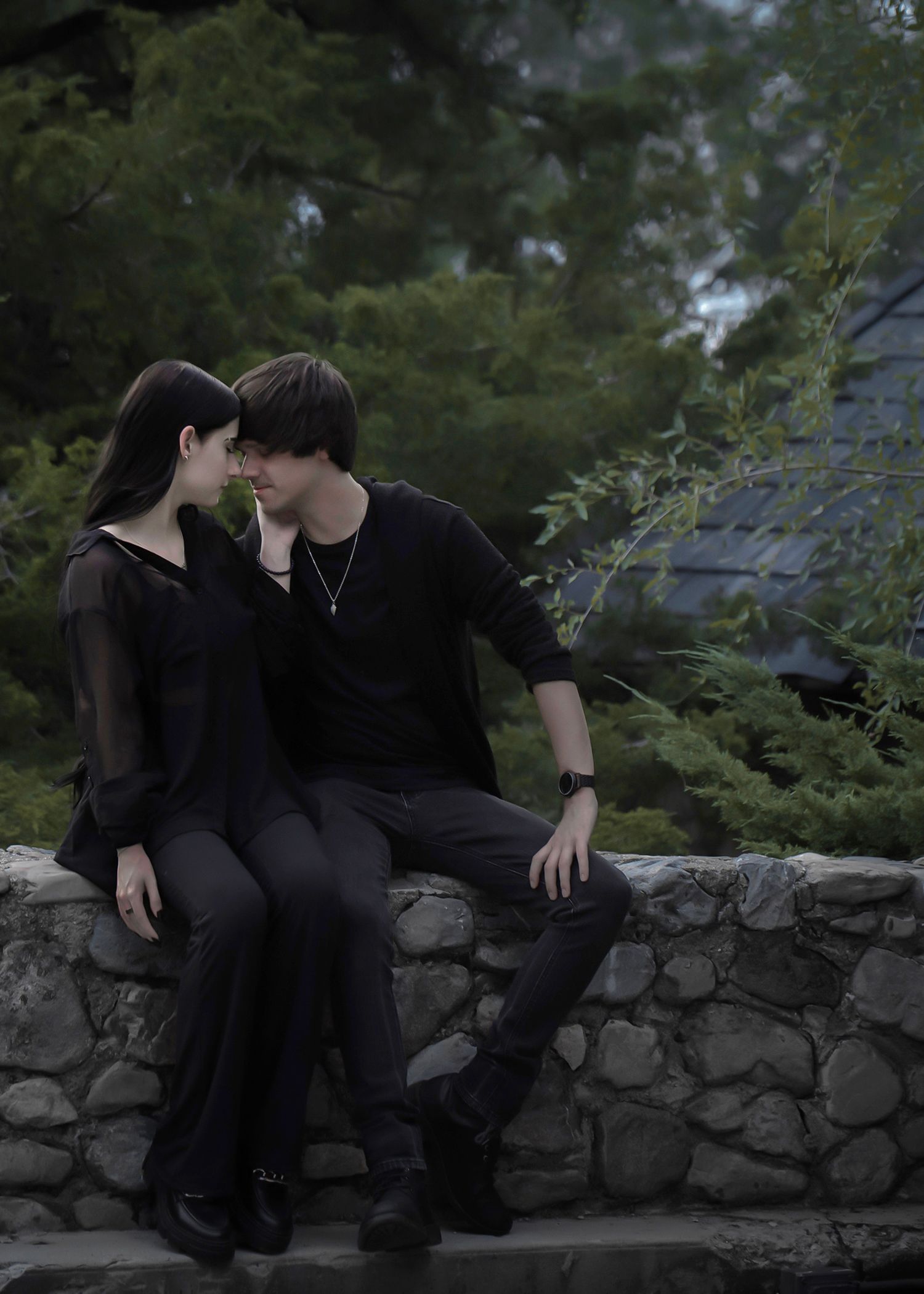 Two people wearing black outfits sit close together on a stone wall surrounded by greenery in a moody outdoor setting.