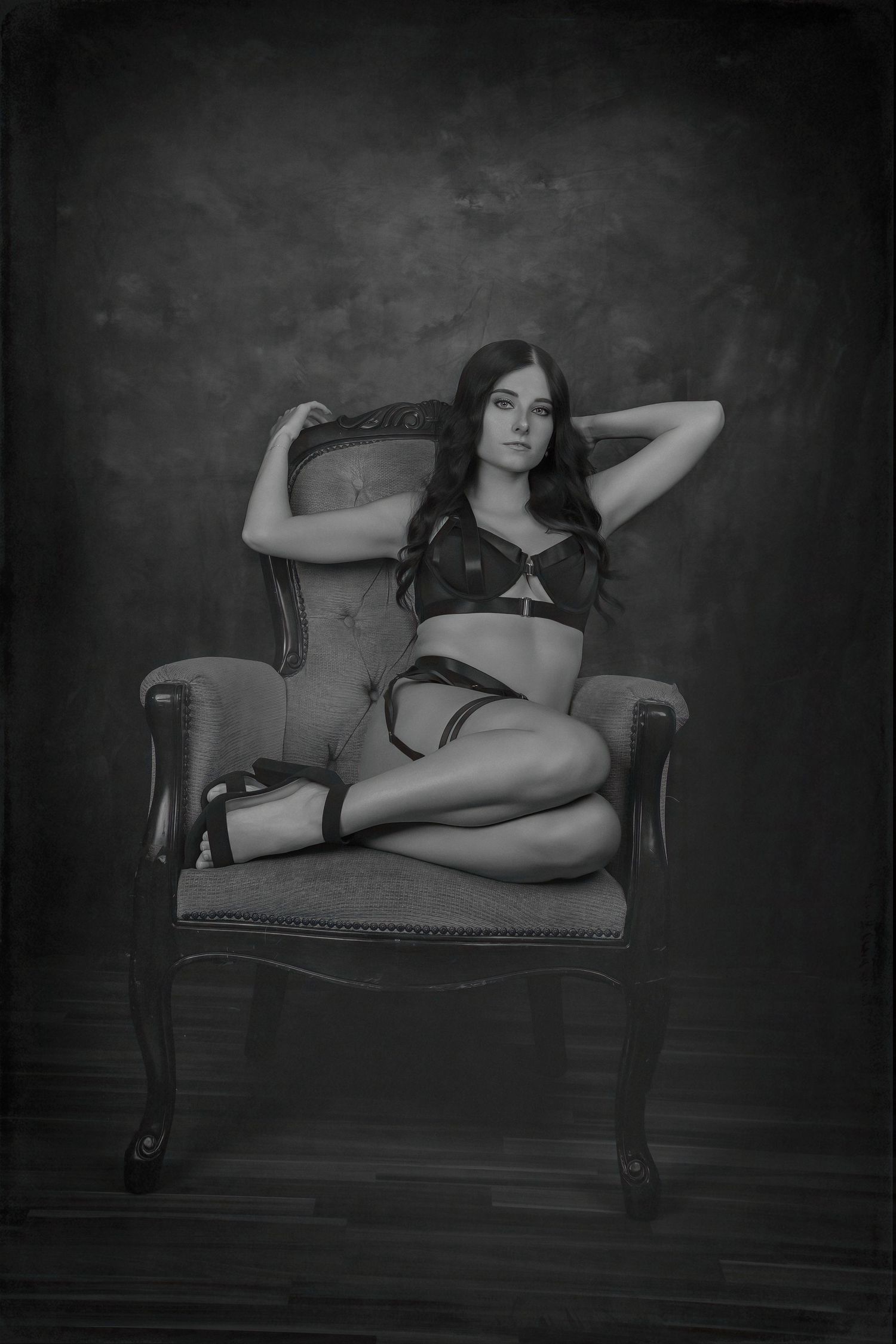 Black and white boudoir photo of a woman in lingerie posing gracefully on a vintage armchair in a studio.