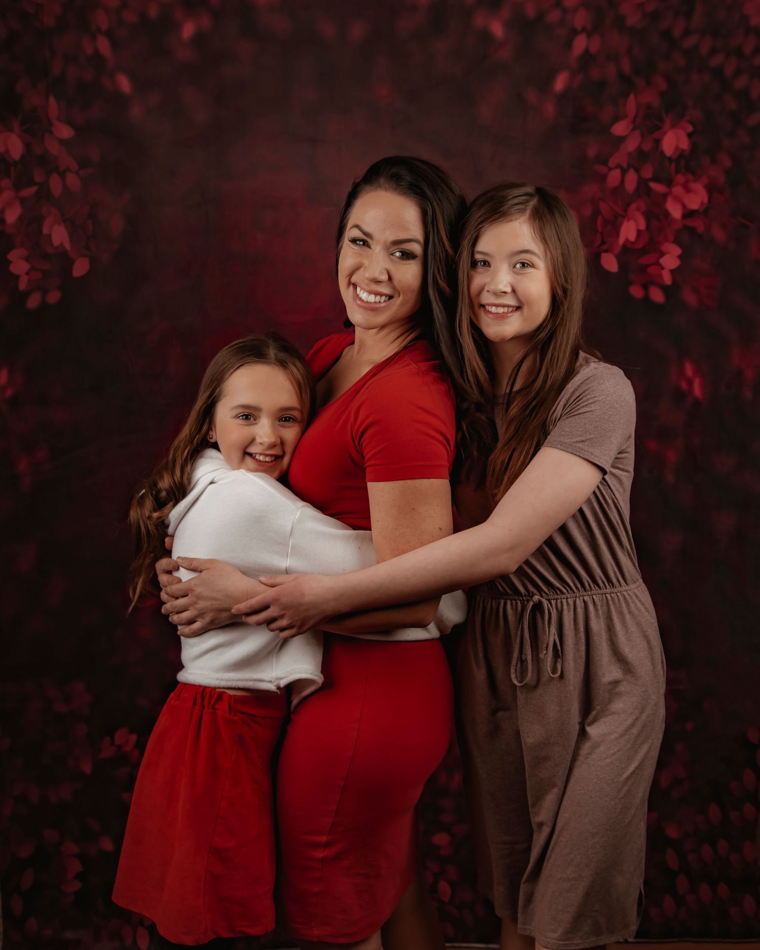 Three people in coordinating red and brown outfits embrace against a dark floral backdrop in a studio portrait.