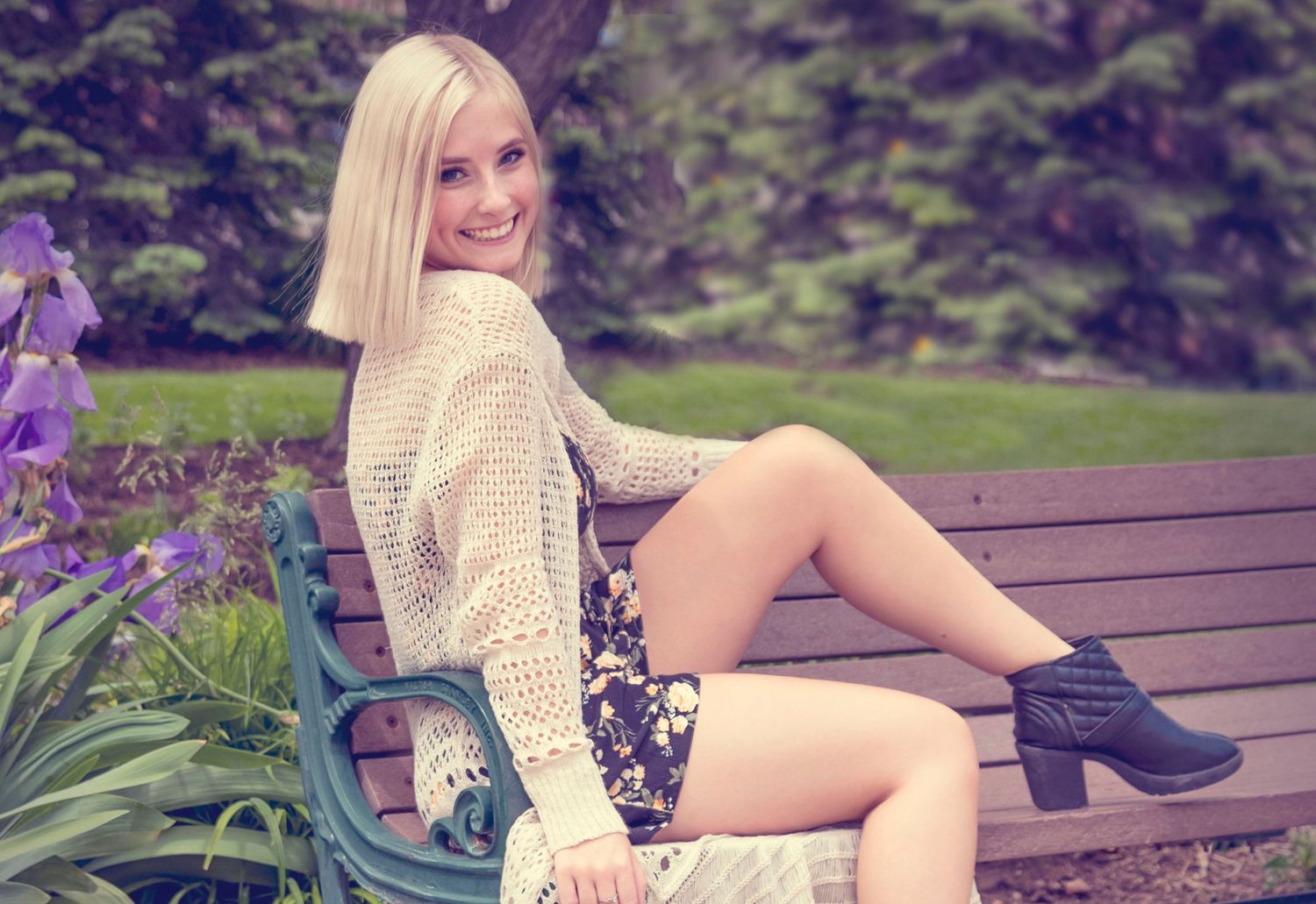 Person in cream sweater and floral dress sitting on garden bench near purple flowers.