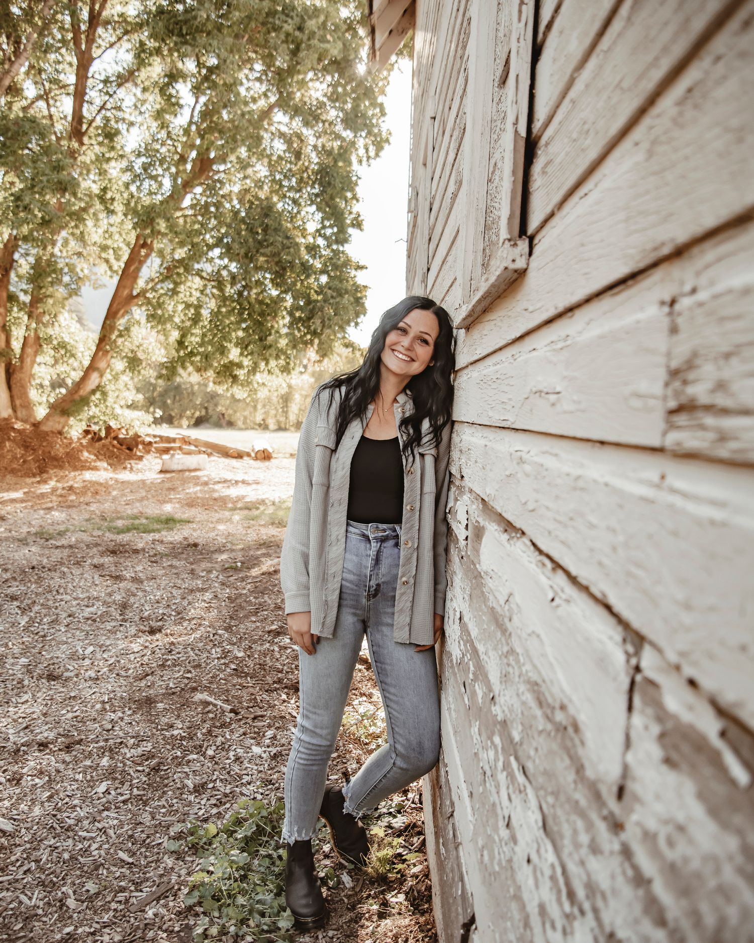 A woman in casual attire leaning against a white wooden wall in warm sunlight.