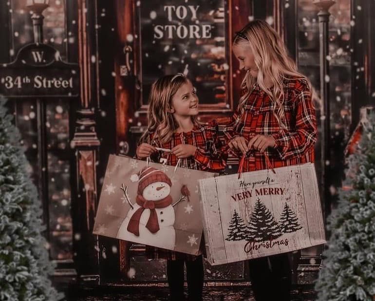 Two people hold holiday shopping bags outside a toy store decorated with Christmas lights and evergreen branches.
