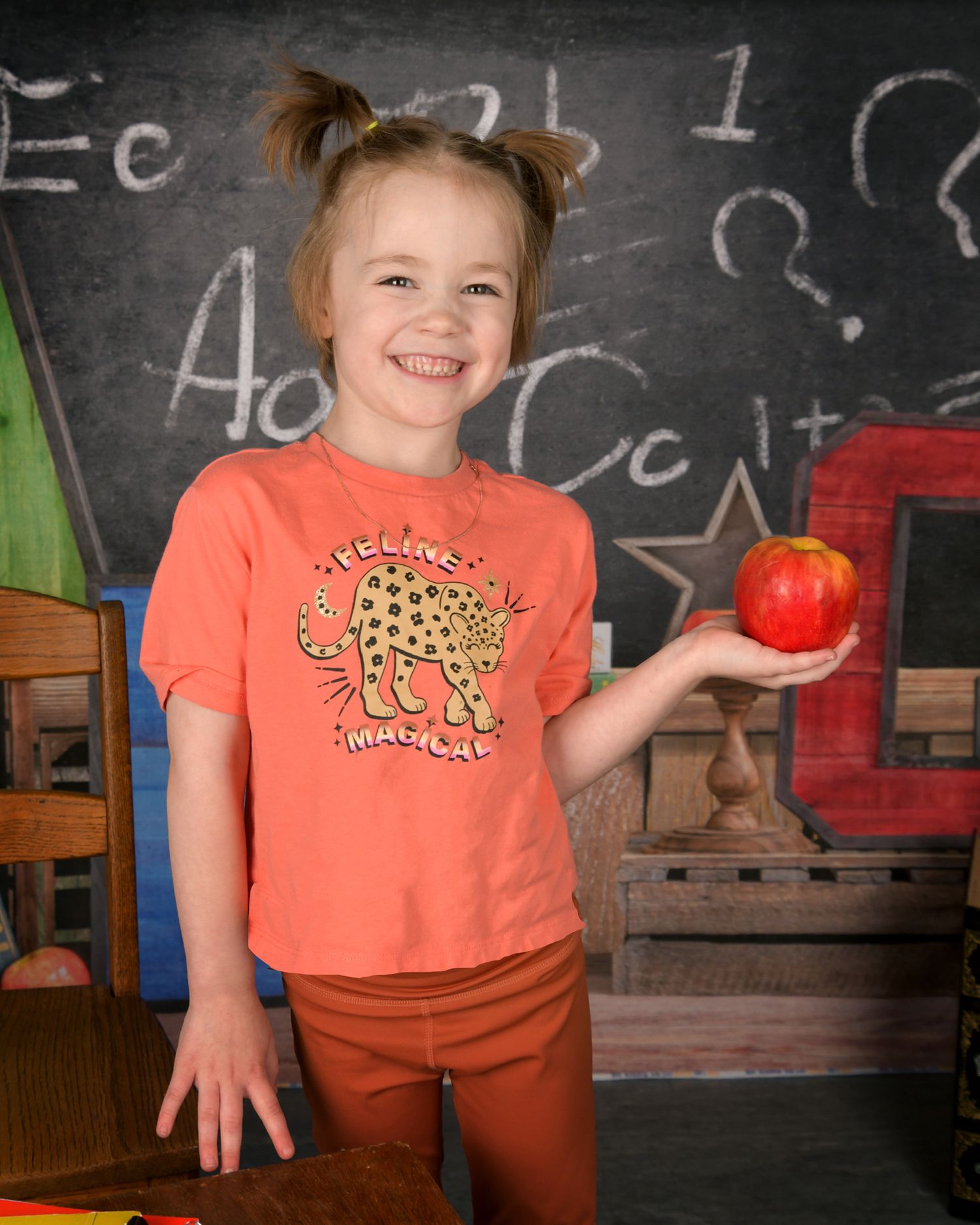 A child in an orange shirt holds a bright red apple while standing at a chalkboard in a classroom setting.
