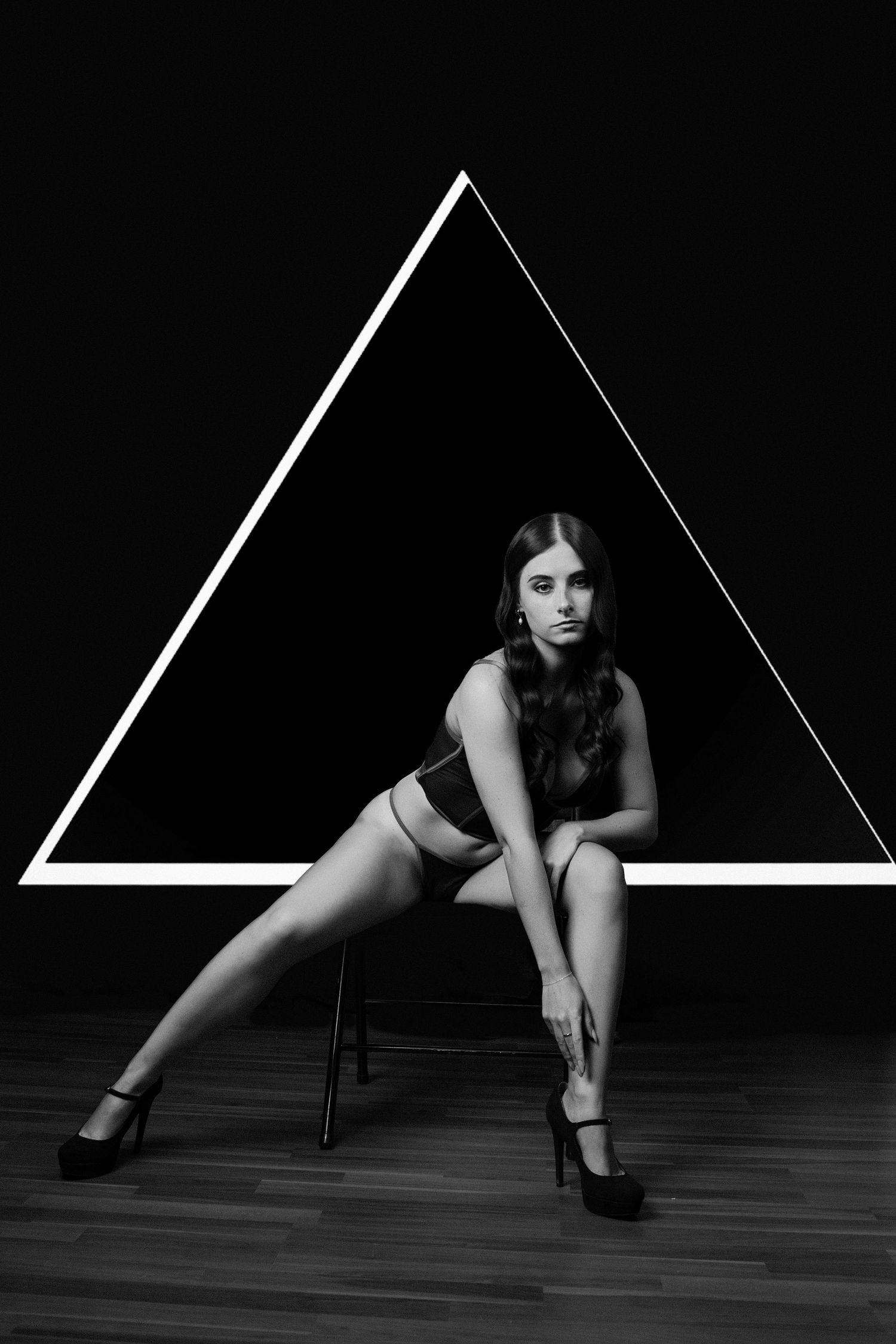 Black and white photo of a woman in lingerie sitting on a chair in front of a glowing triangle on a dark background.