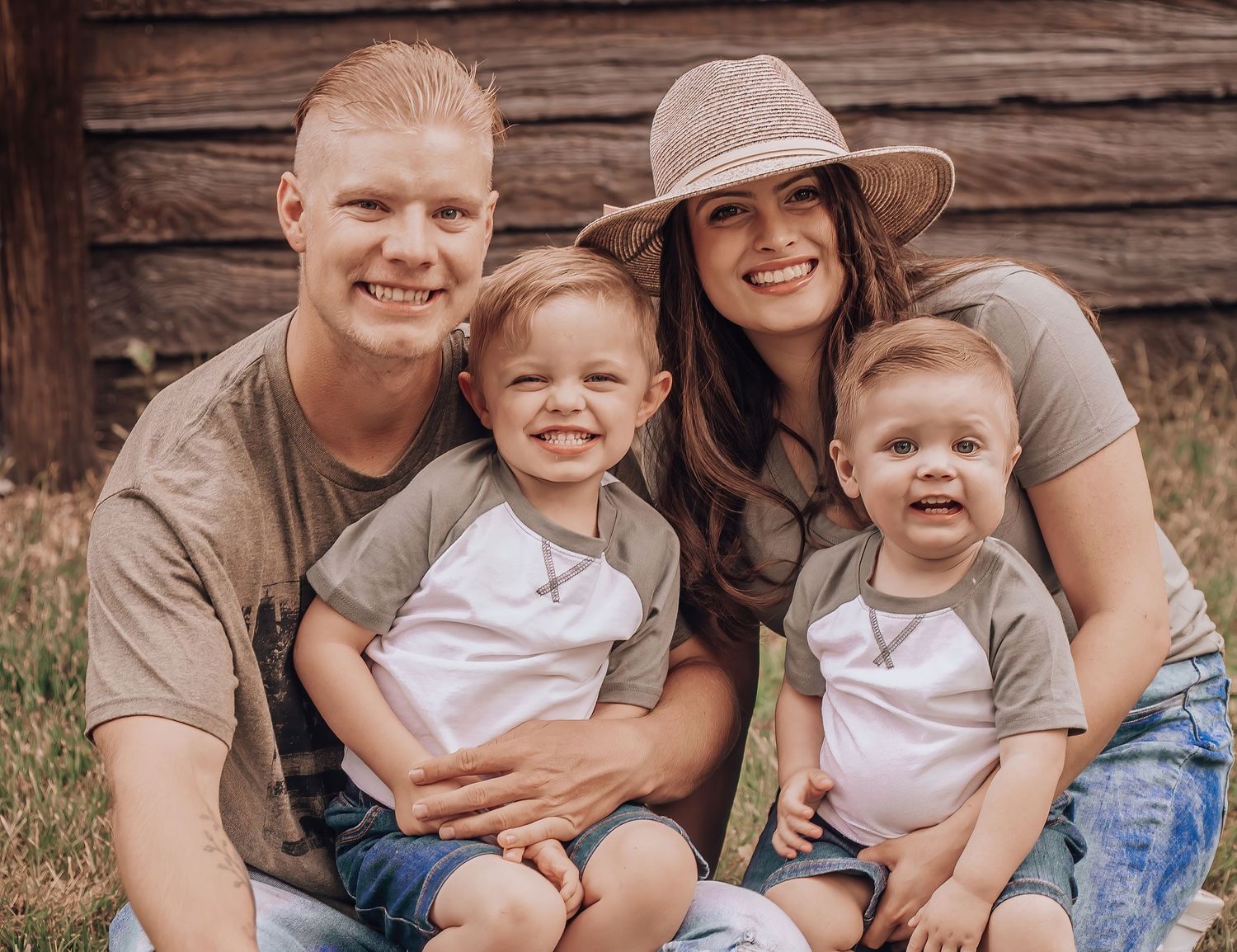 Family of four sits together against rustic wooden wall wearing coordinated neutral colors.