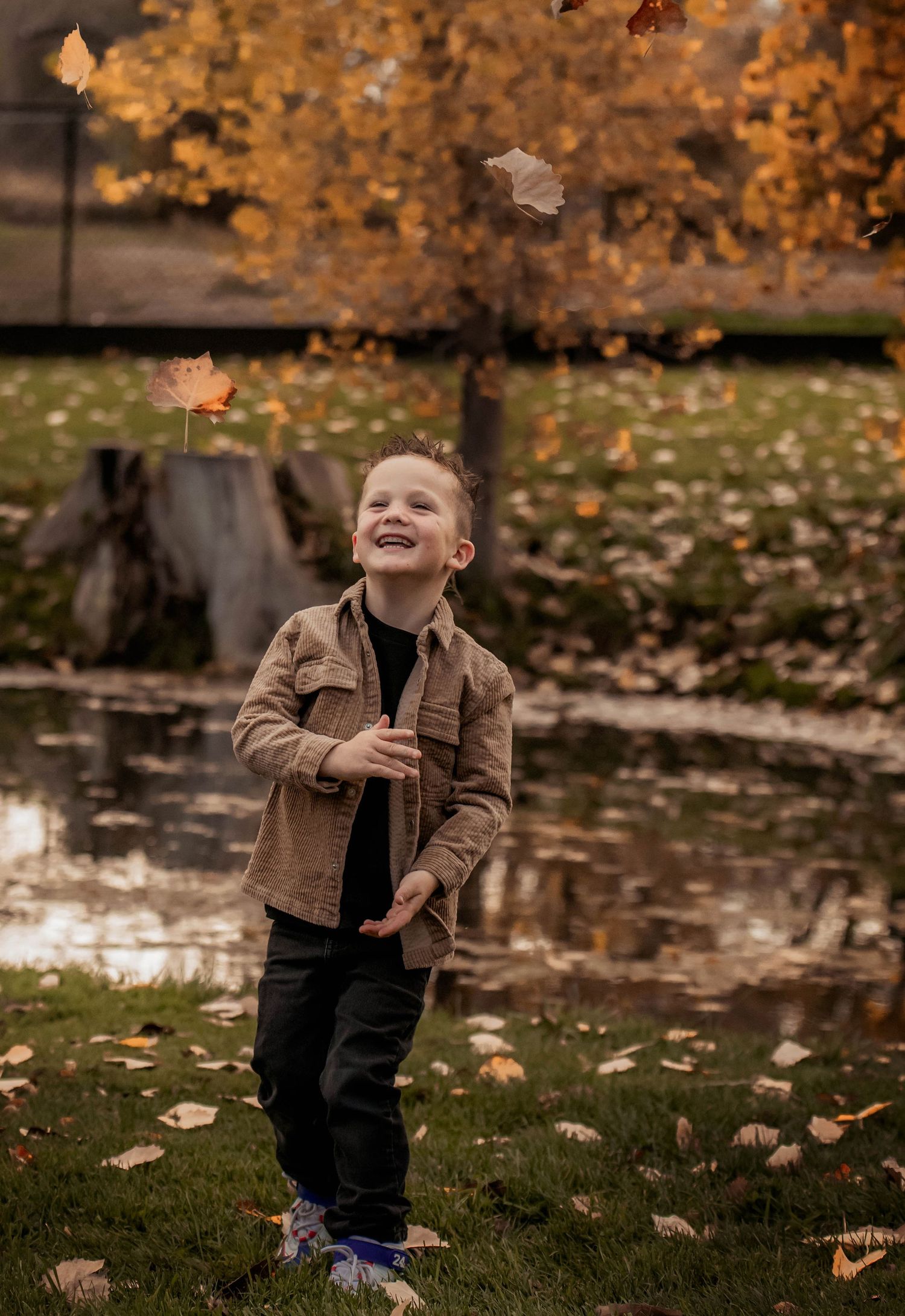 Child playing outdoors in autumn with falling leaves wearing brown jacket and dark pants on grassy ground.
