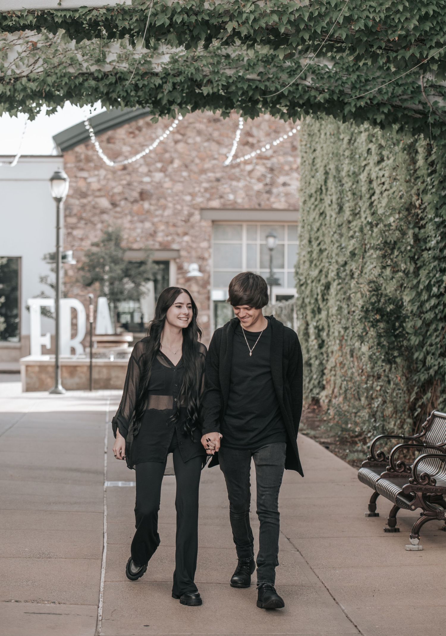 Two people dressed in all black stroll along a brick sidewalk holding hands near a stone building.