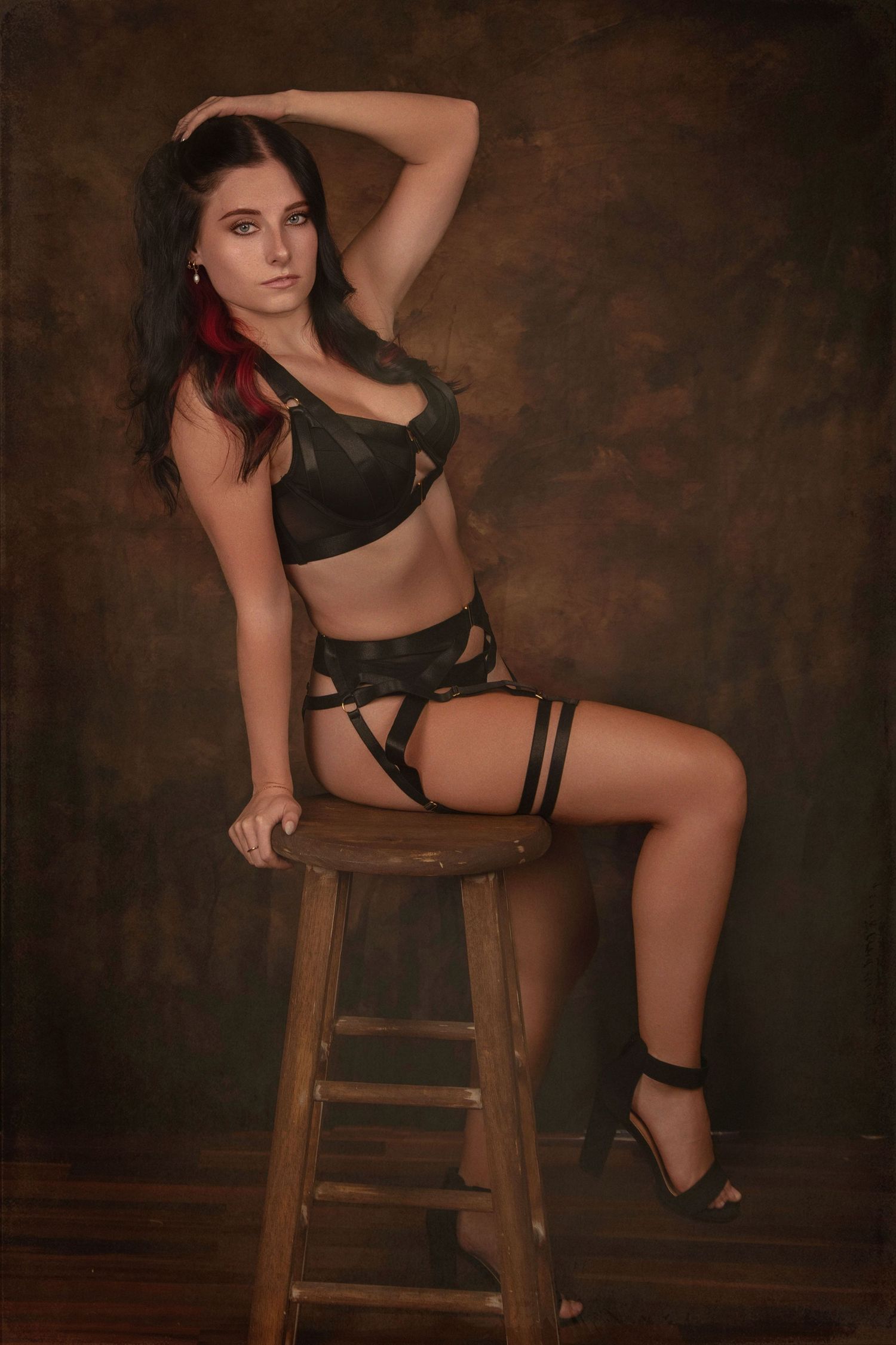 Woman in dark lingerie and strappy garter belt seated on wooden stool against brown textured studio backdrop.