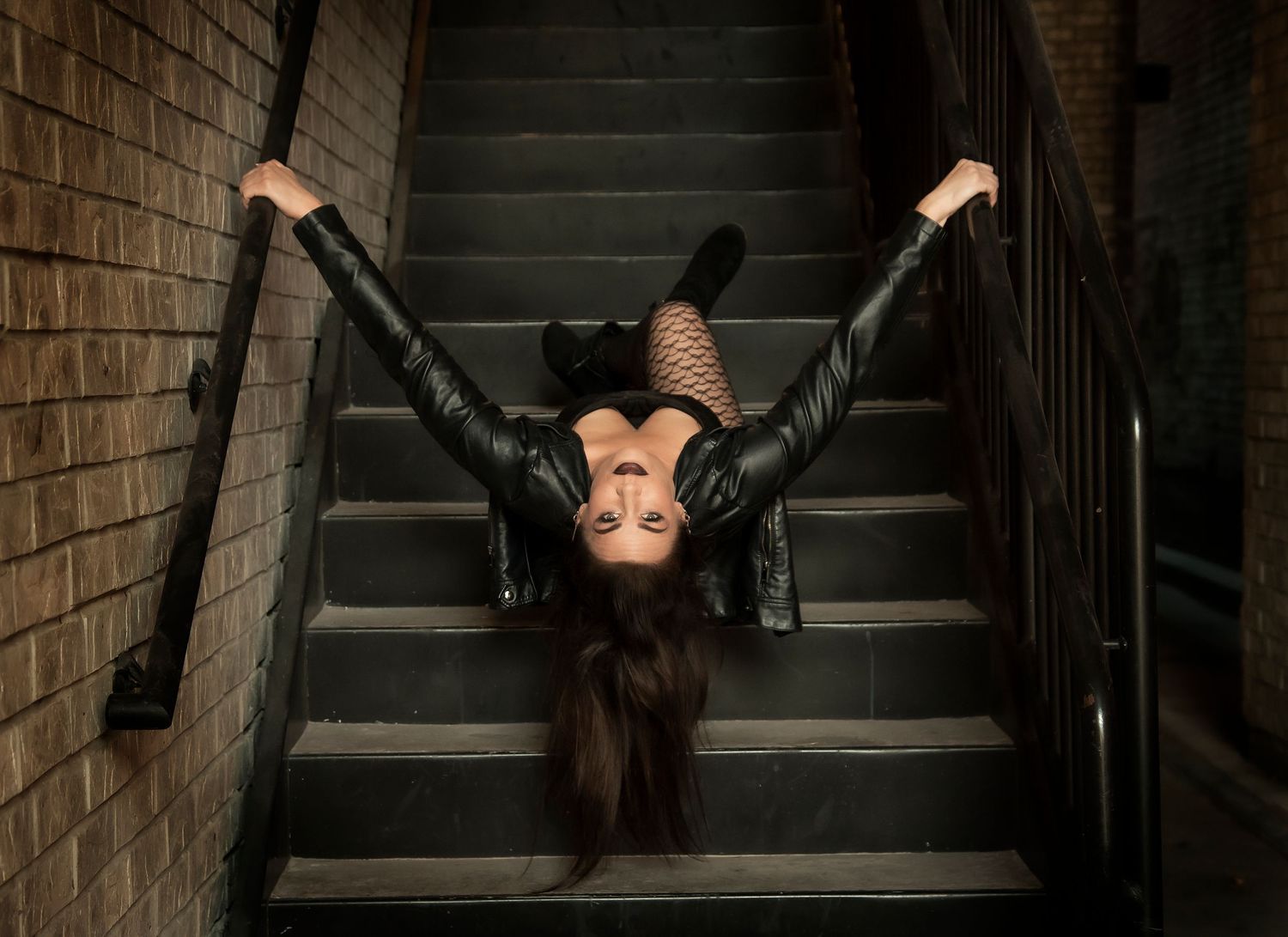Dramatic upside down pose on dark staircase with moody lighting and leather outfit.
