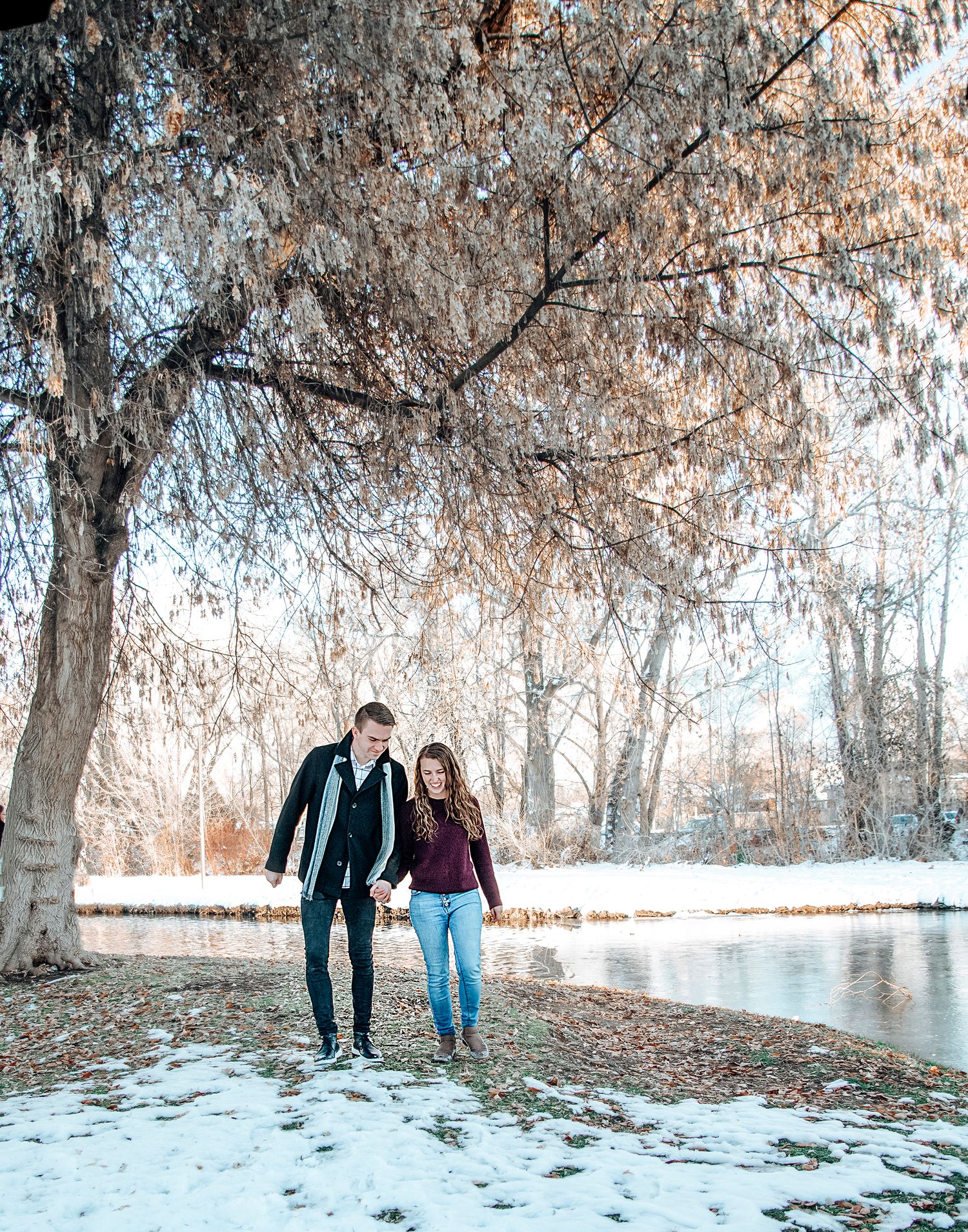 A couple strolls together along a snowy riverbank beneath frost-covered trees.