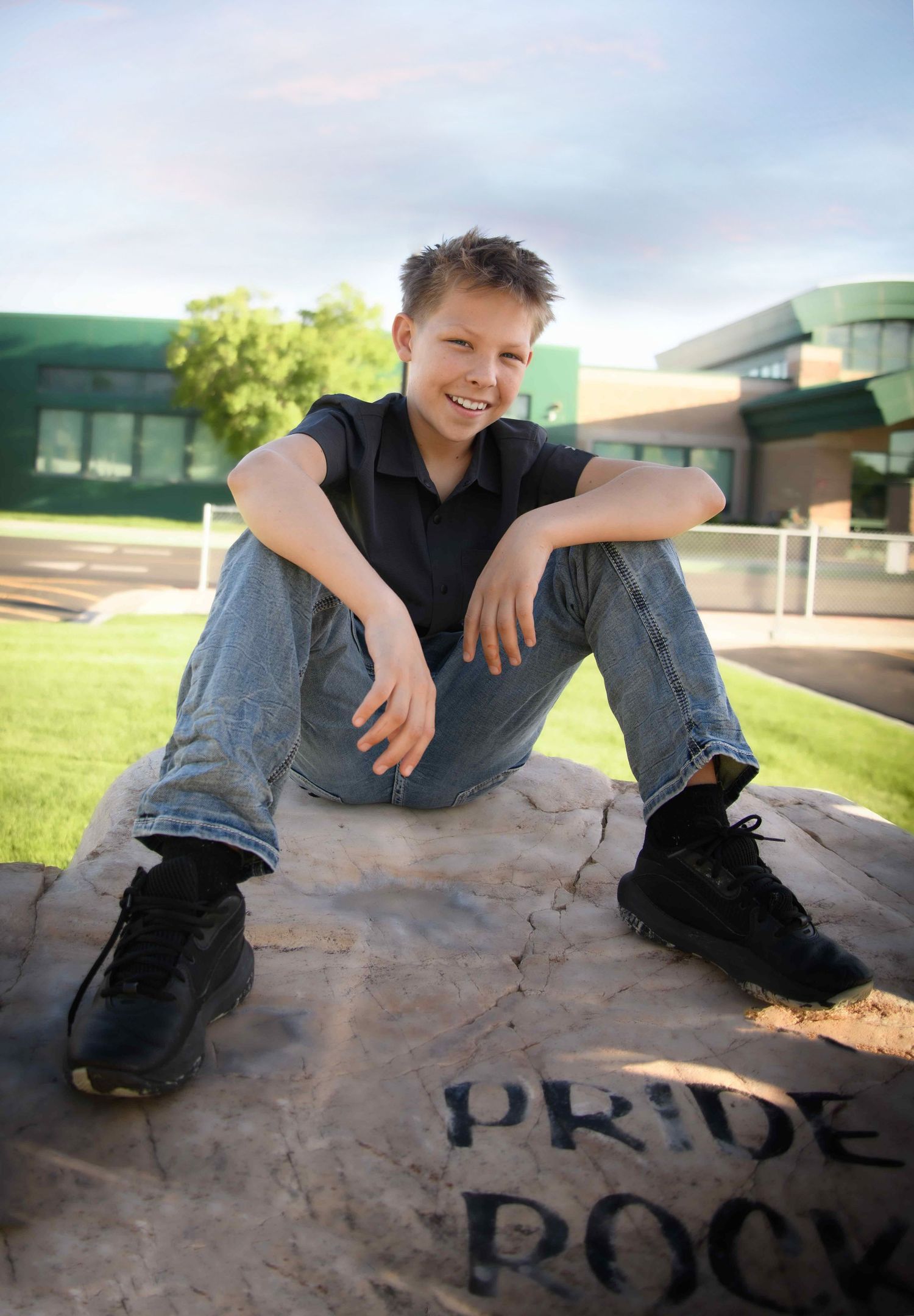Smiling teenage boy sitting on Pride Rock outside a school building on a sunny day.