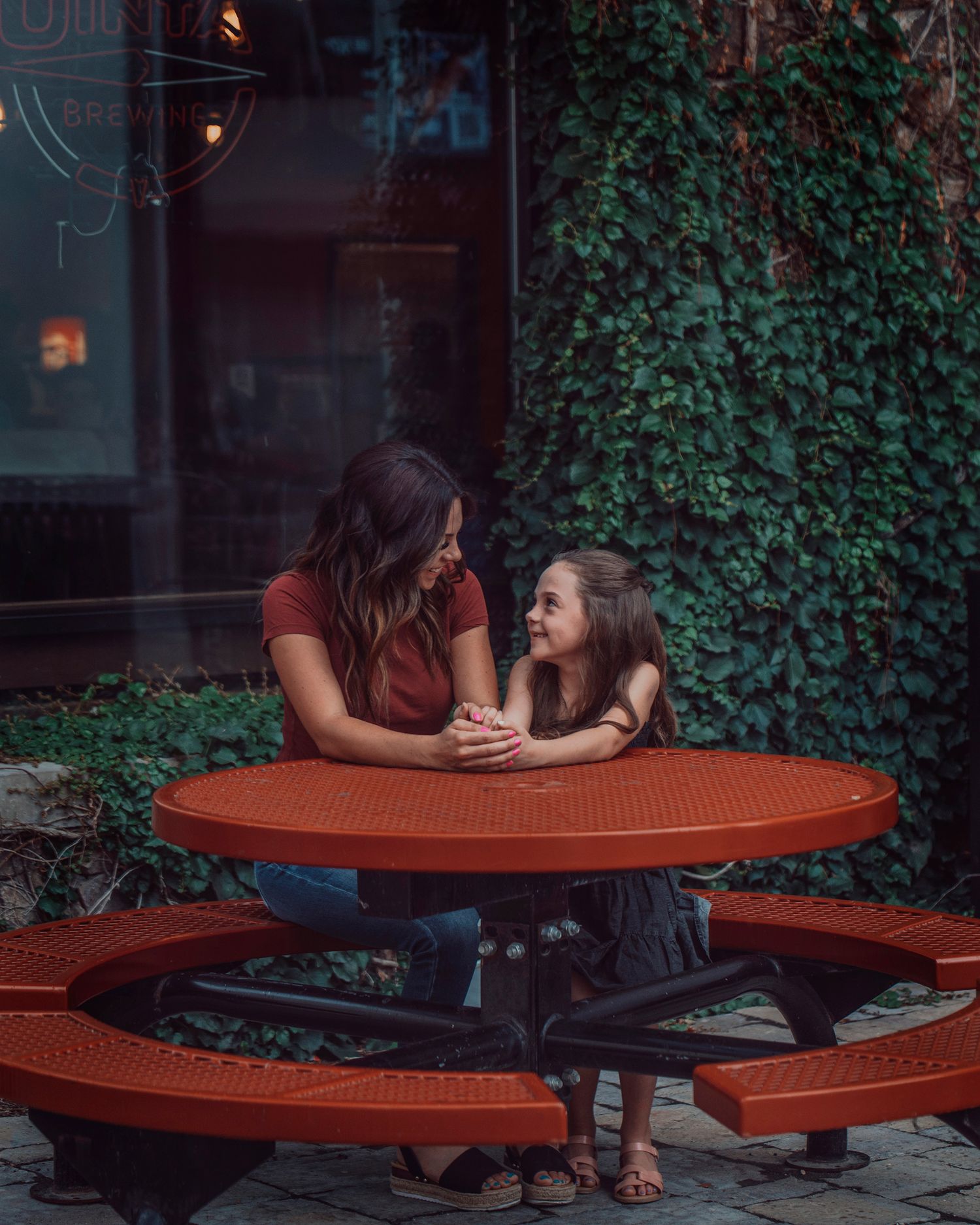 Two people sitting at red outdoor picnic table surrounded by ivy-covered walls.