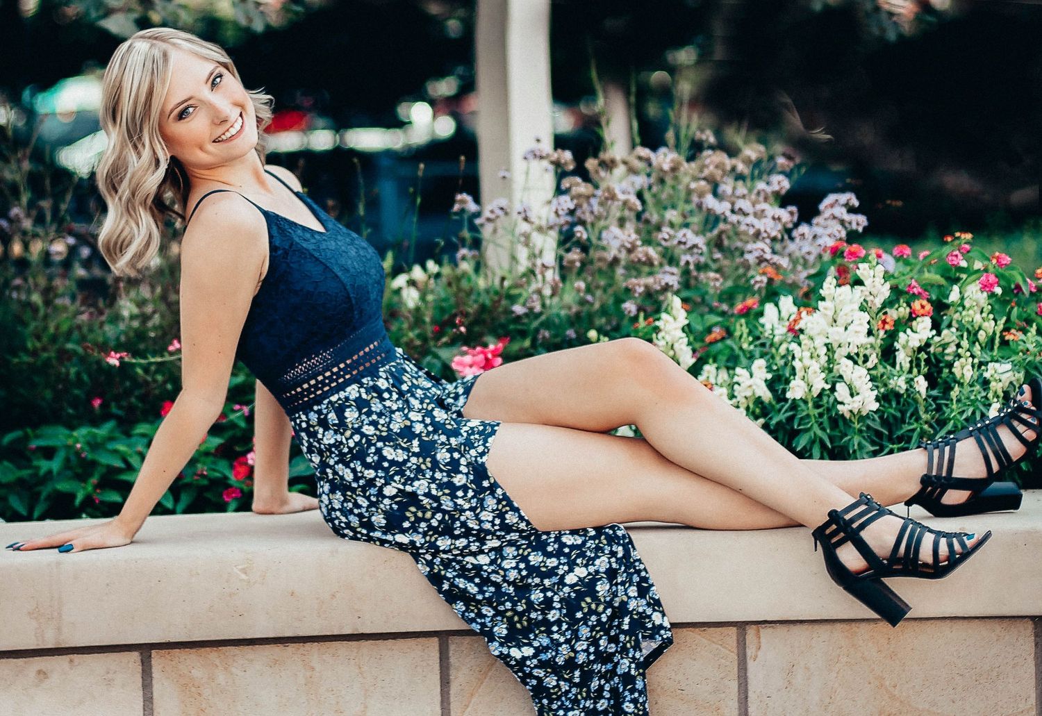 Person in a blue dress posing on a stone wall near a garden with flowers.