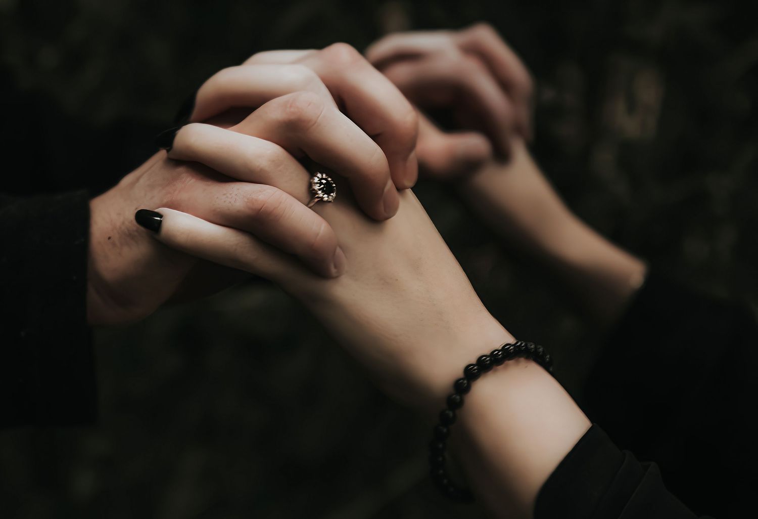 Close-up shot of hands with black nail polish and jewelry against a dark background.
